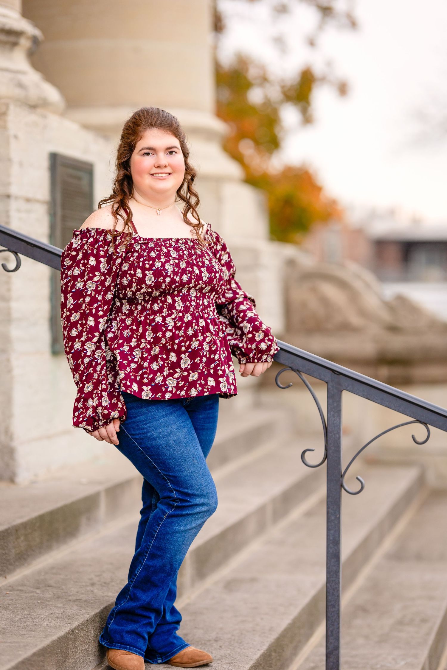 Woman in floral burgundy top poses on concrete steps with iron railing during autumn.