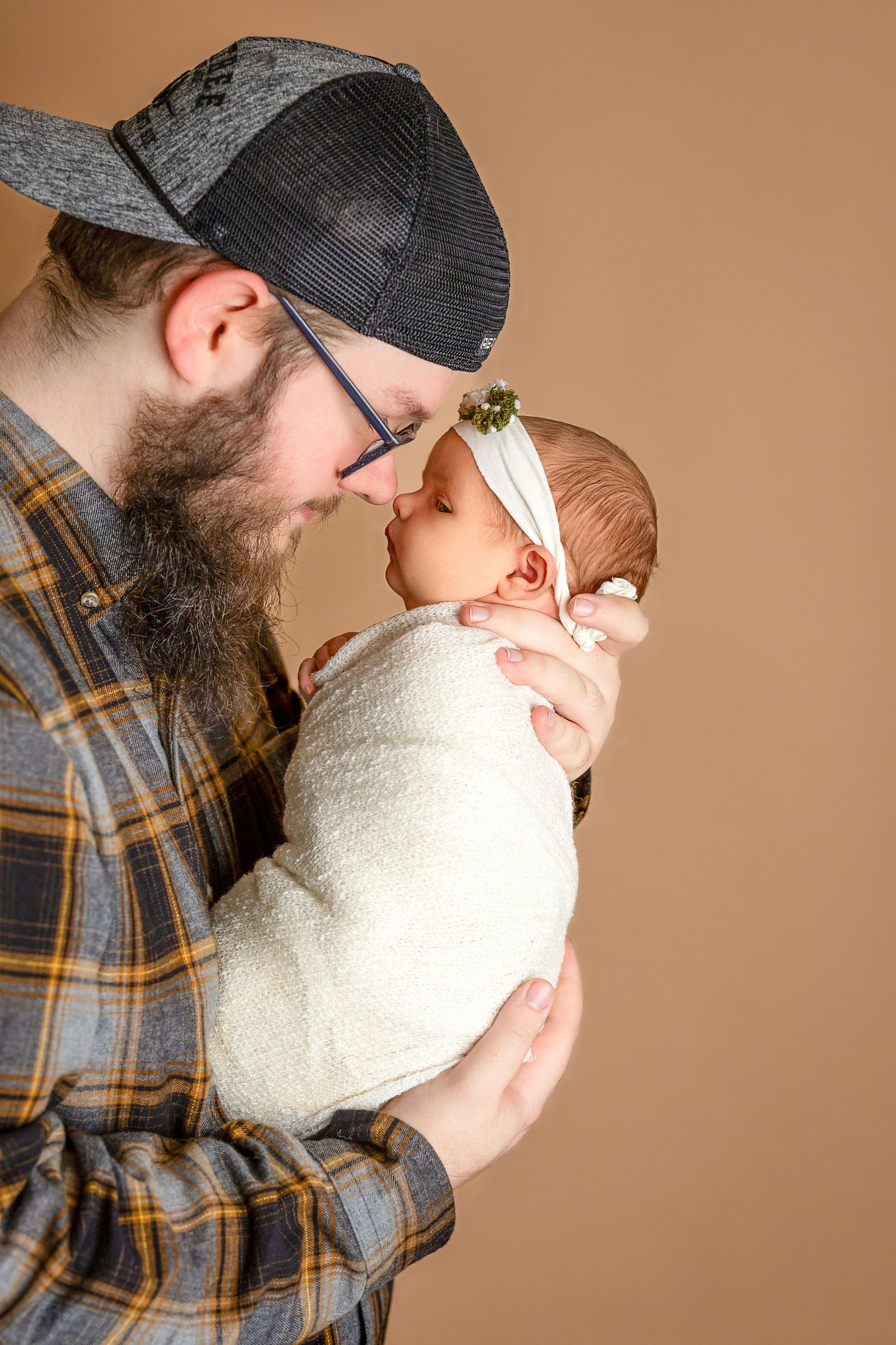 A sweet moment between a swaddled newborn and caregiver wearing a plaid shirt and baseball cap.