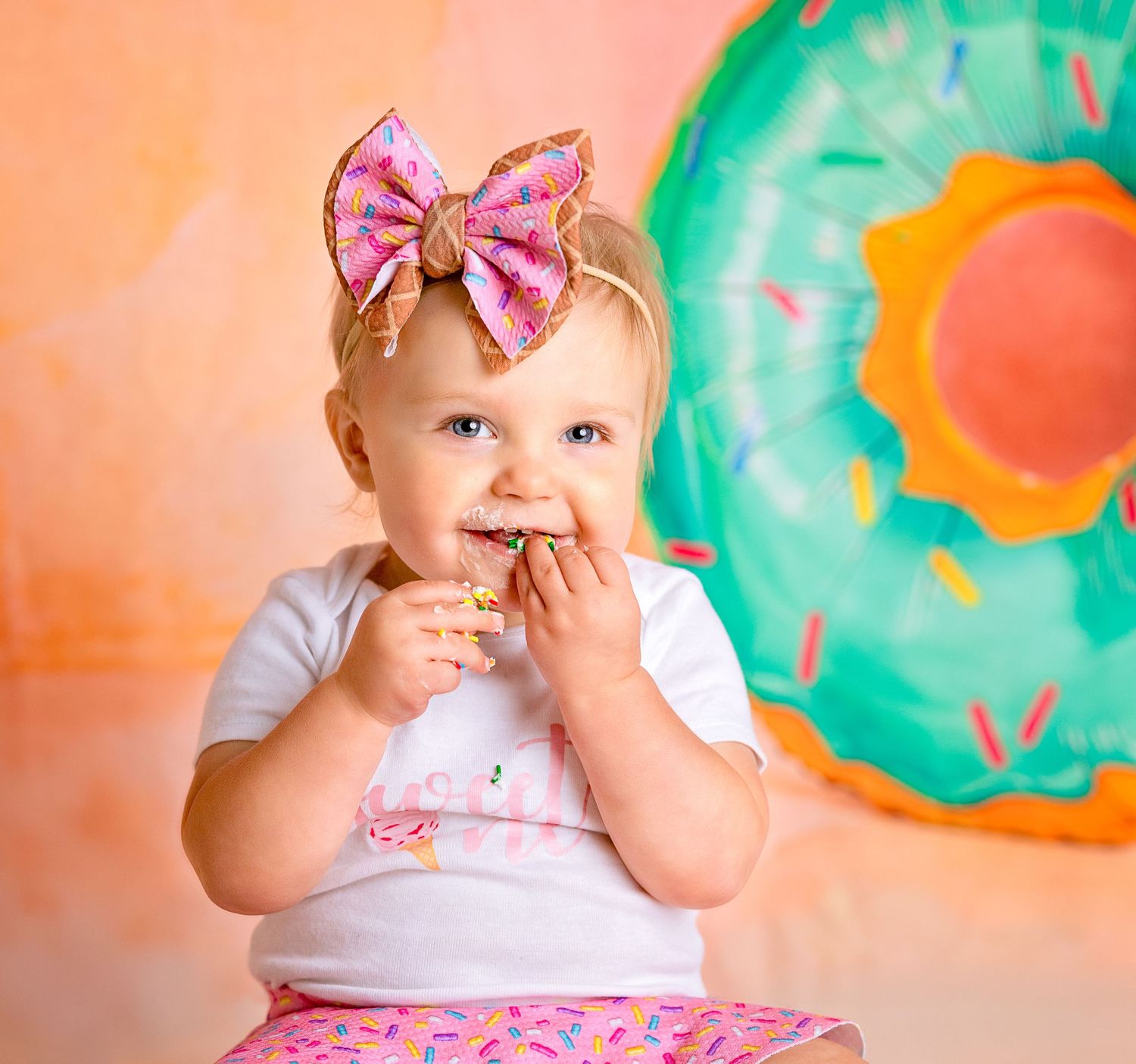 Baby in pink bow wearing tutu smiles while eating cake with colorful donut backdrop.
