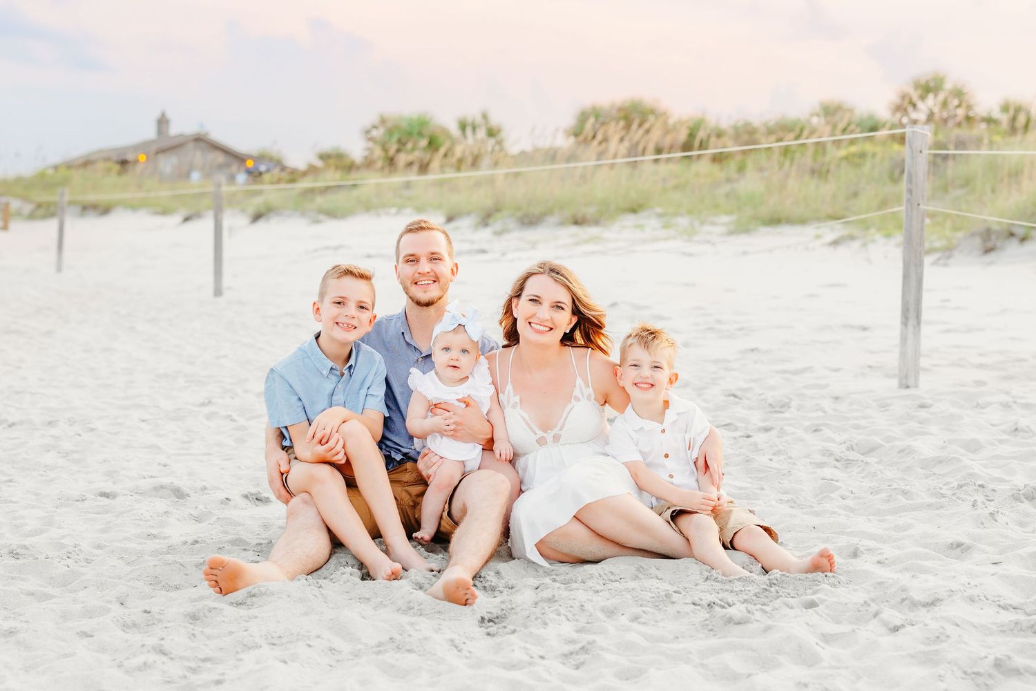 A family sits together on a sandy beach with dunes and fencing in the background during a sunny day.