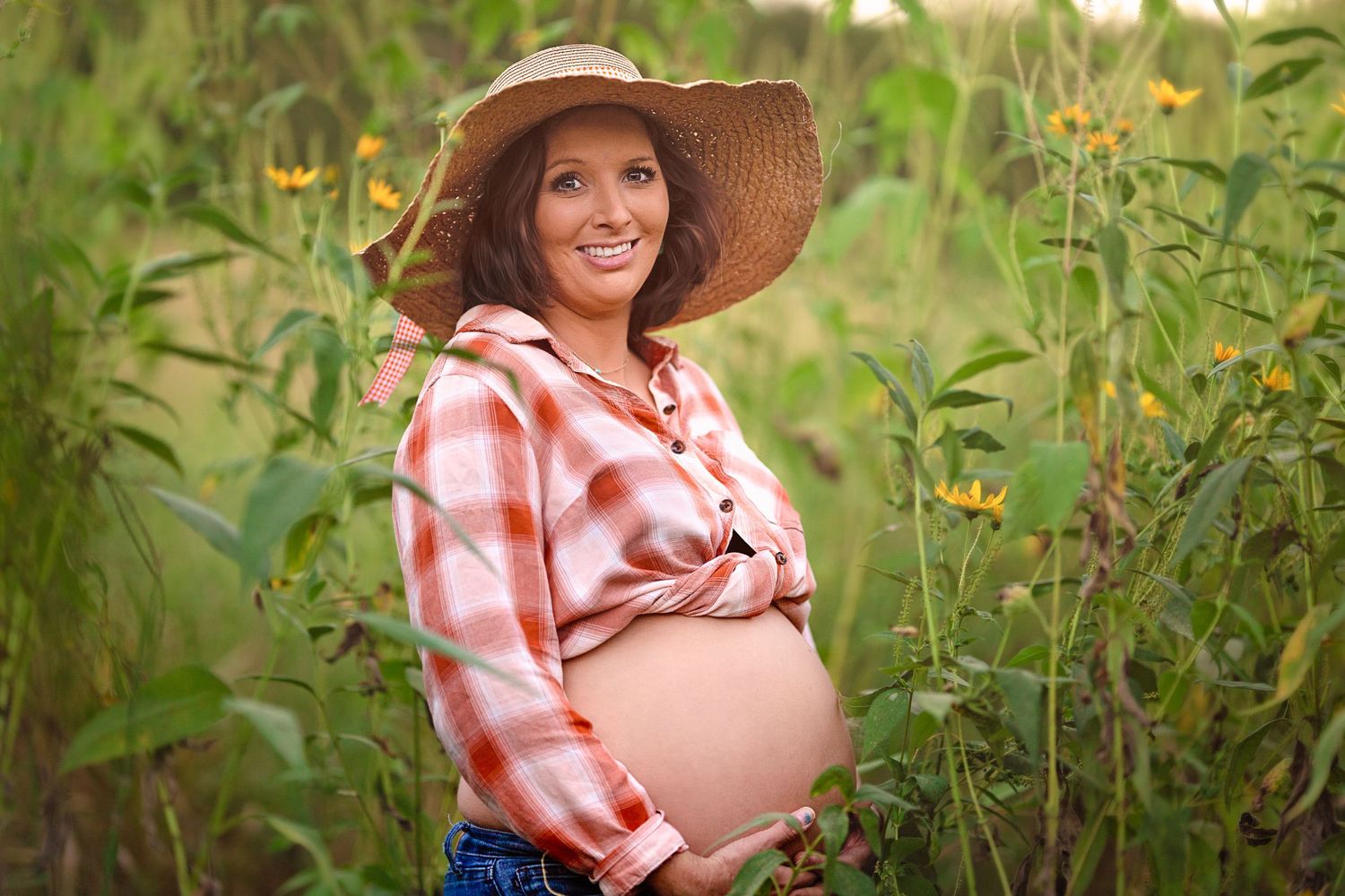 Maternity photo of expectant mother in a sunhat and plaid shirt standing among sunflowers.