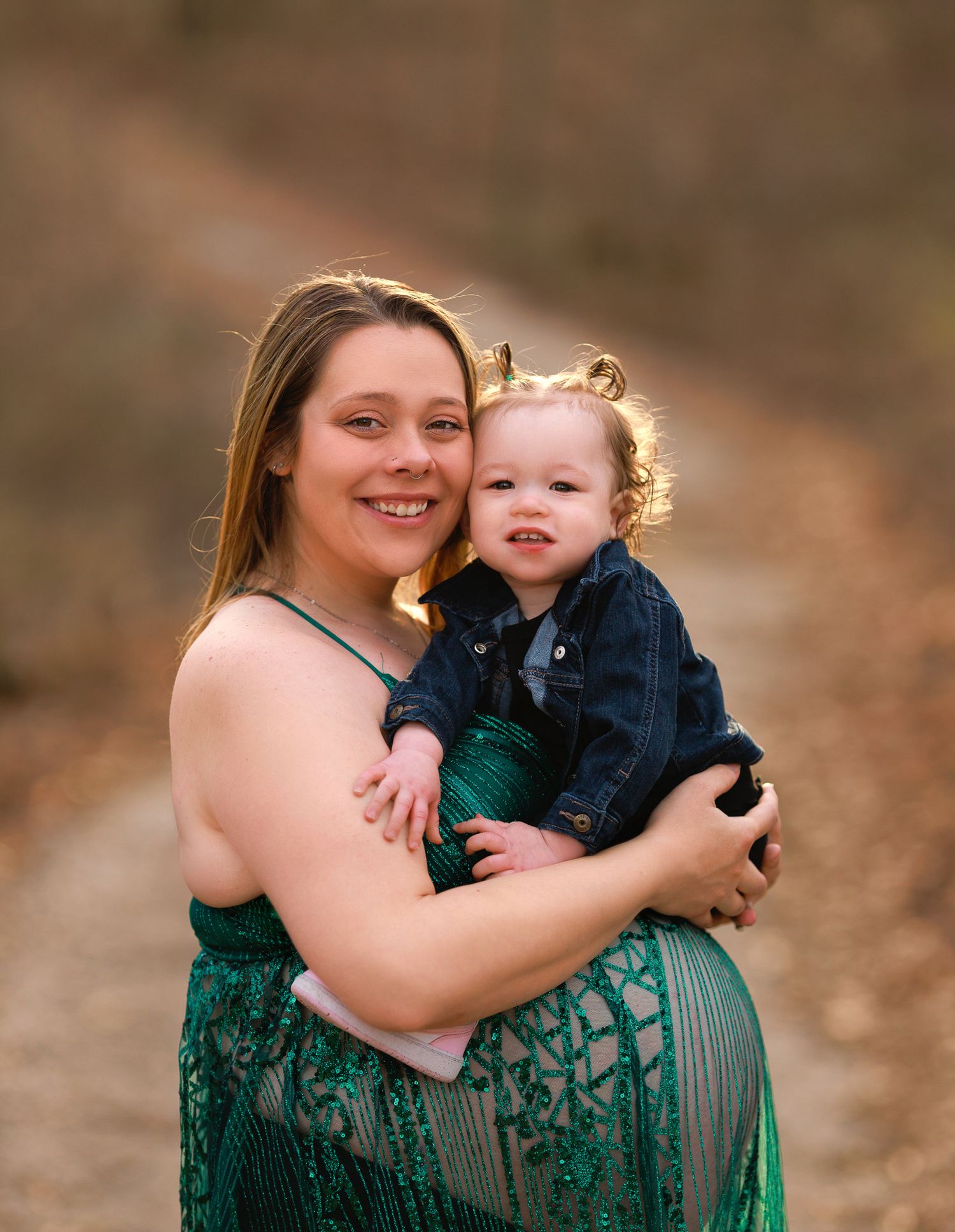 A maternity portrait of a woman in a green lace dress holding a small child outdoors.
