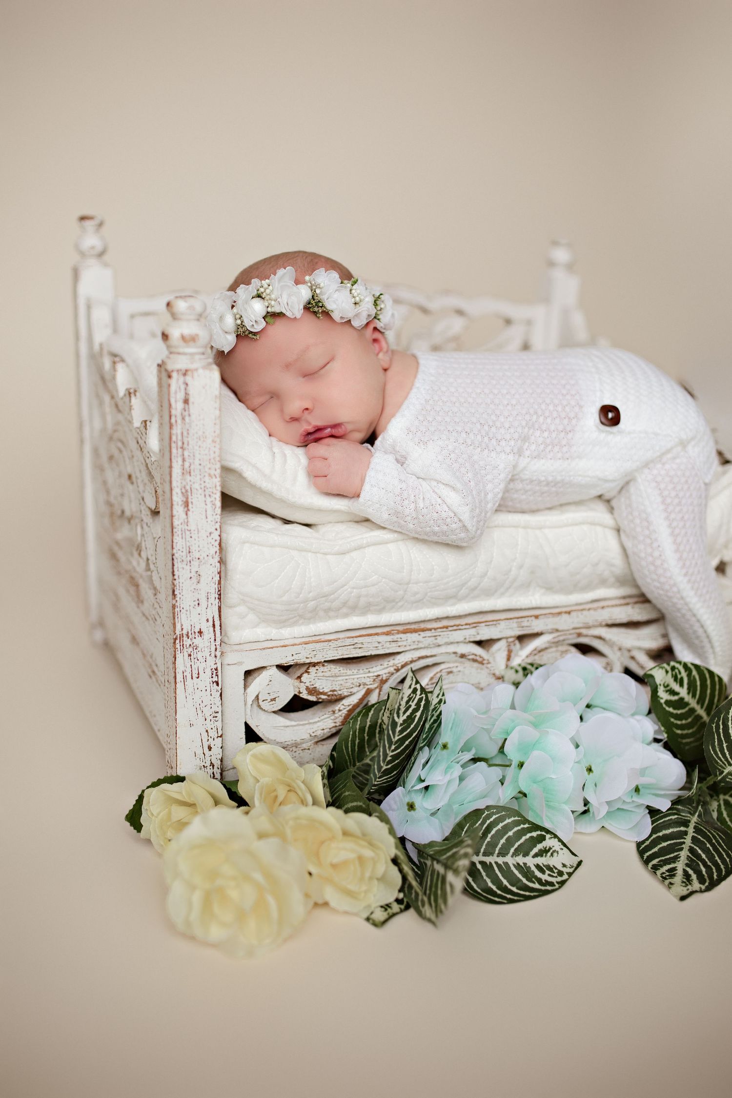 Newborn sleeping peacefully in vintage white bed with floral crown and white knit outfit.