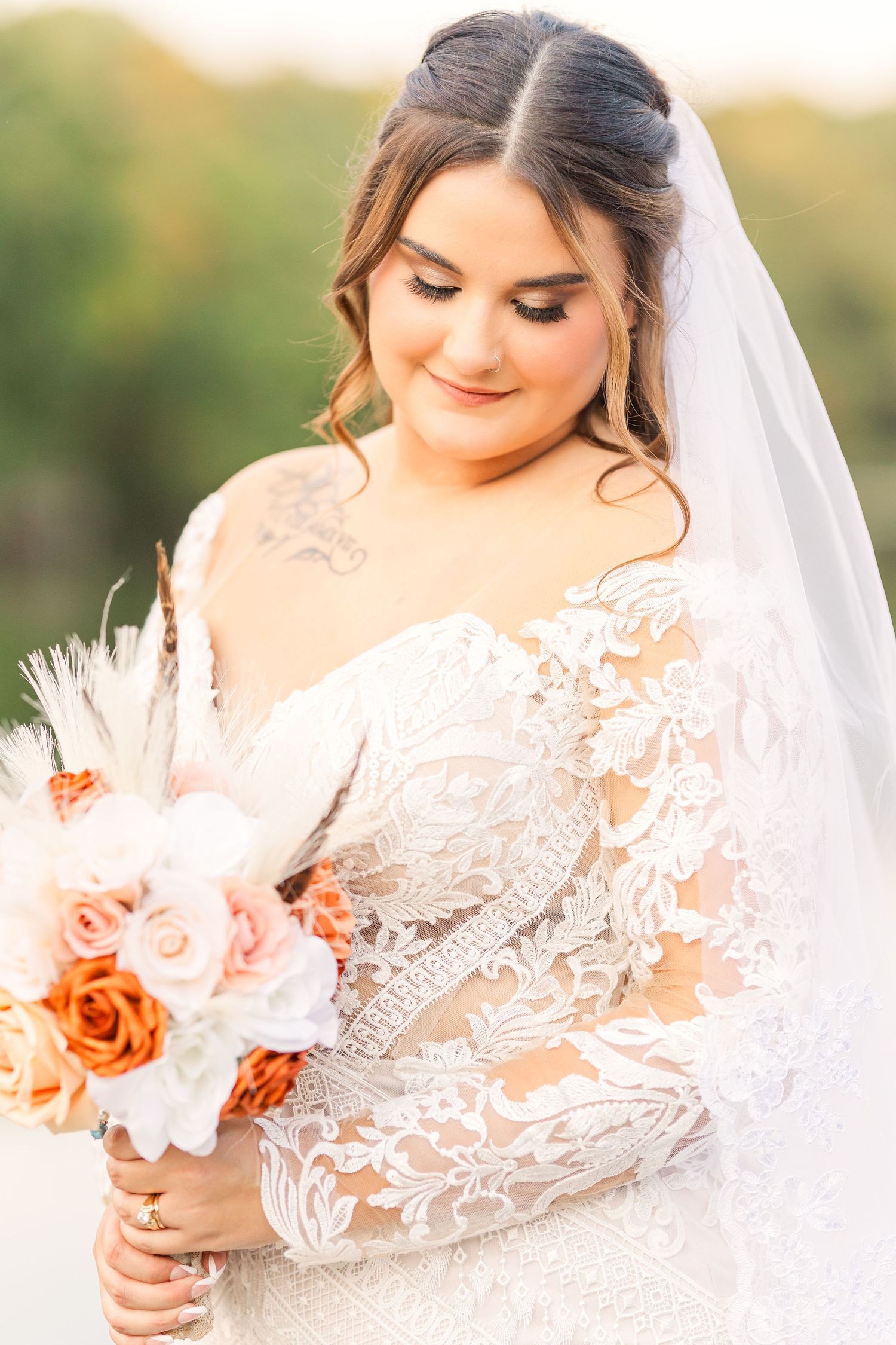 A bride in a lace long sleeve wedding dress holds a peach and white bouquet while looking down.