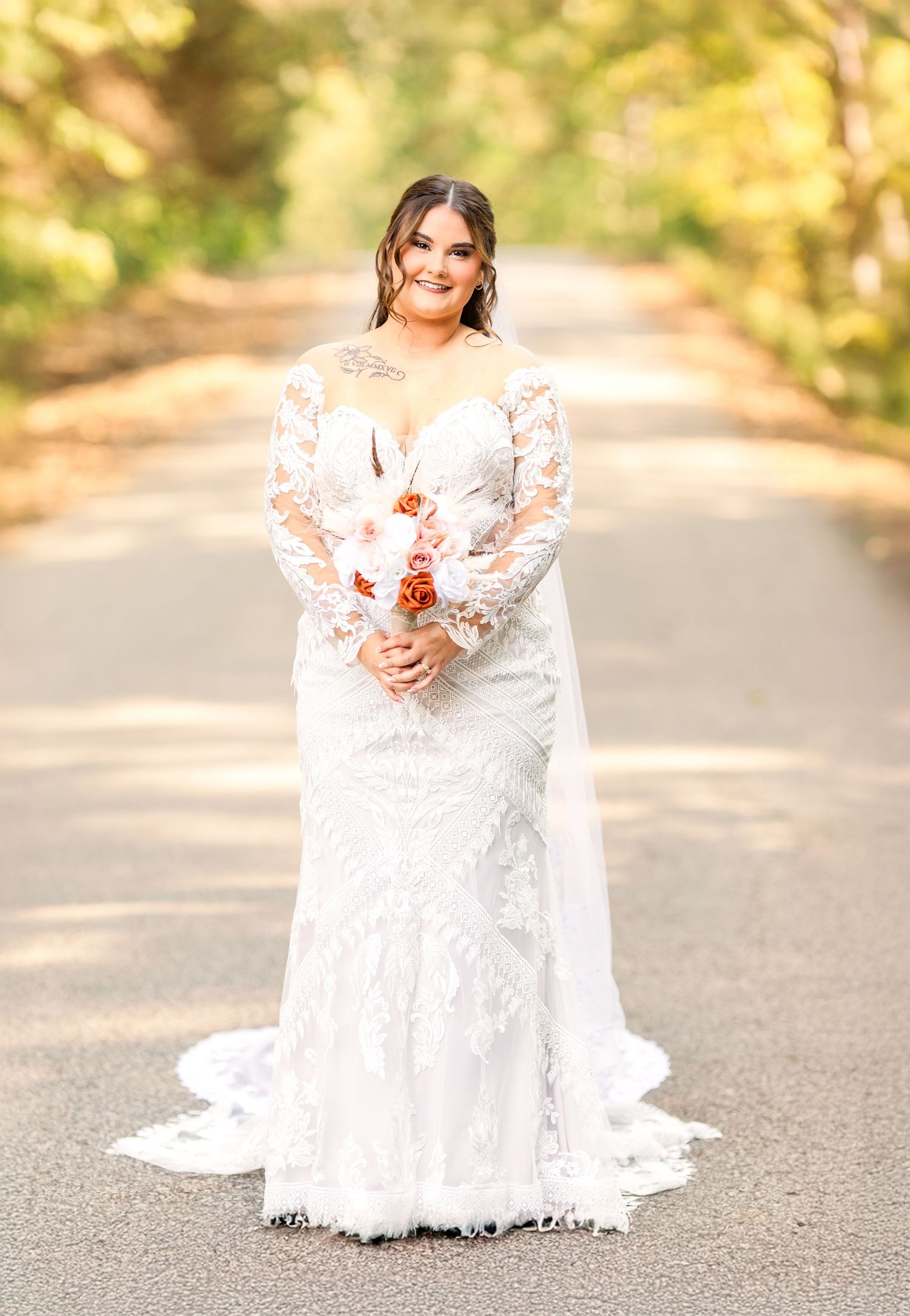 A bride in a lace gown poses on a fall road holding a coral and white bouquet.