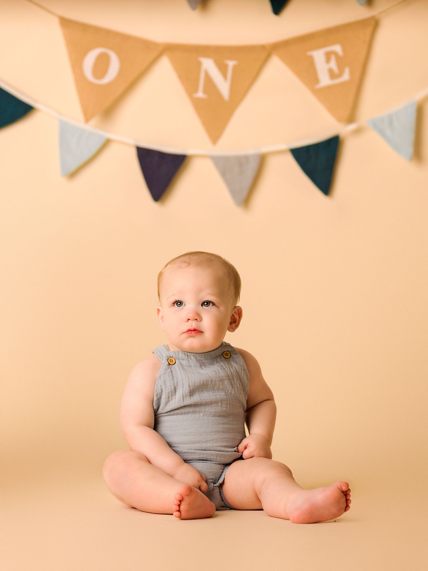 Baby in grey onesie sitting under 'ONE' banner during first birthday photoshoot.