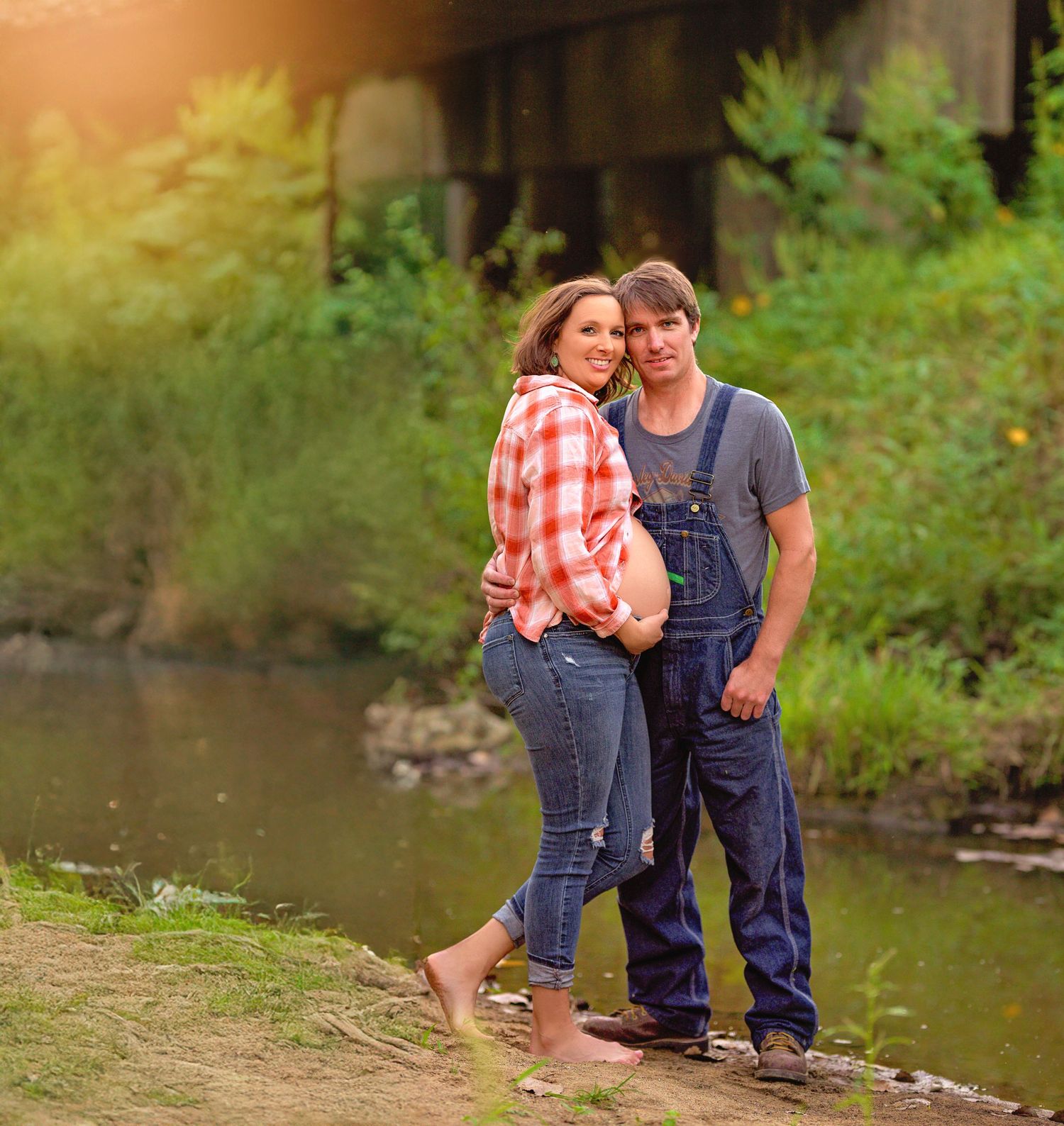 Couple in overalls and plaid stands barefoot by a creek during sunset.