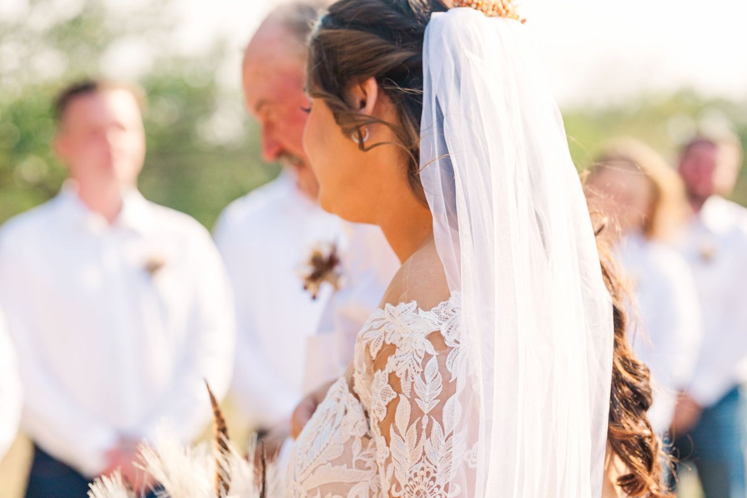 A bride in a delicate lace wedding dress and flowing veil stands during an outdoor ceremony.