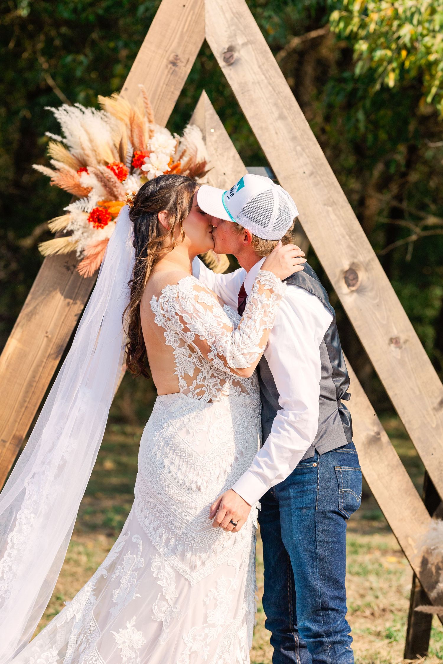 A couple shares a kiss under a wooden triangle arch decorated with dried flowers at their outdoor wedding.