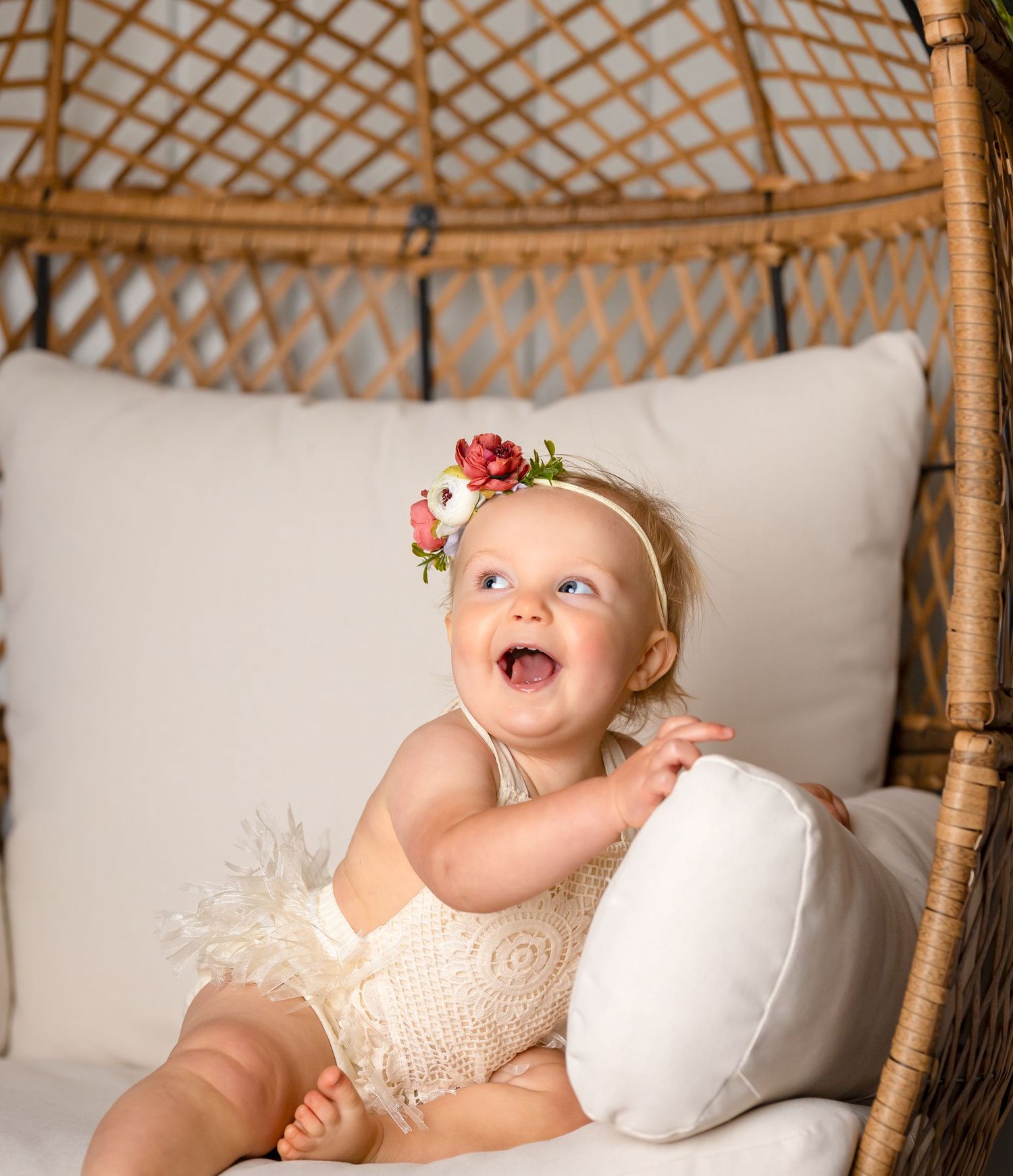 Joyful baby wearing a floral headband sits in a wicker peacock chair with white cushions.