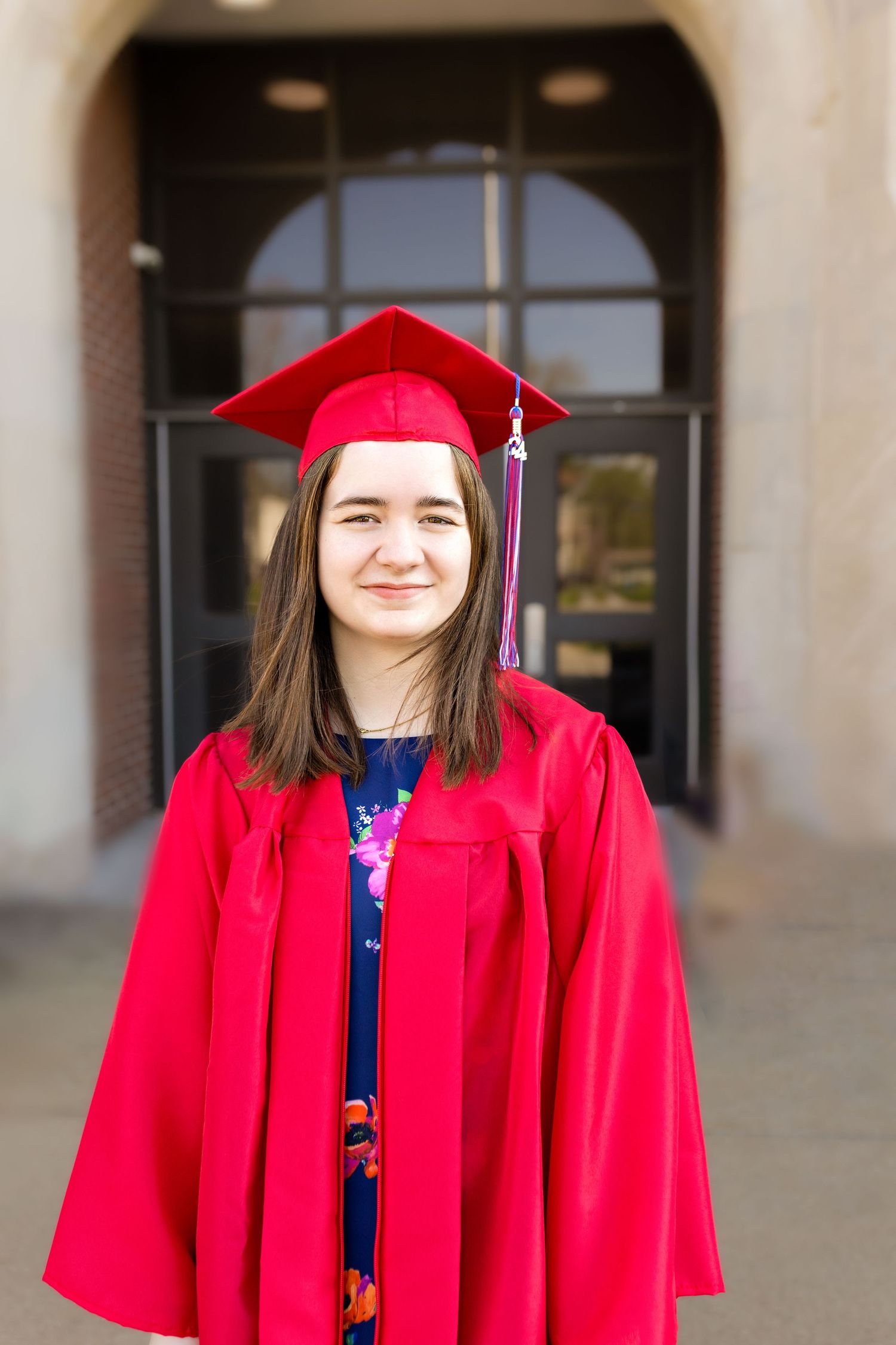 A graduation portrait showing someone in a bright red cap and gown standing in front of an arched doorway.