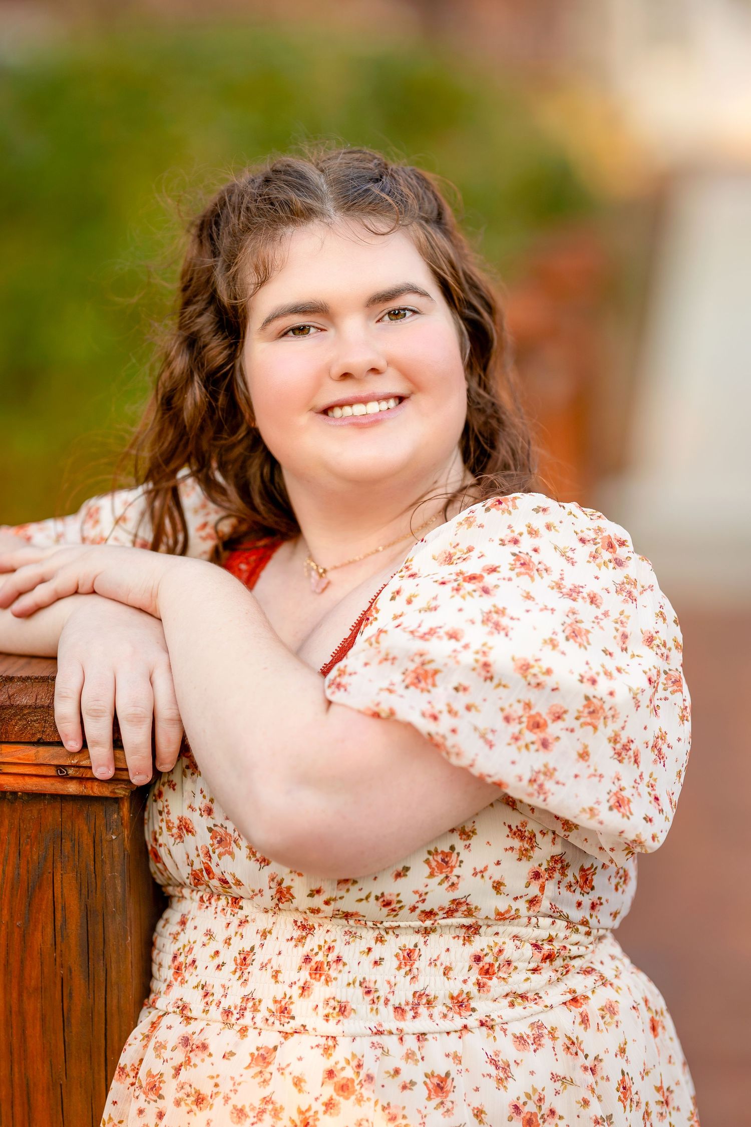 Outdoor portrait in floral dress leaning against wooden railing with warm natural lighting.