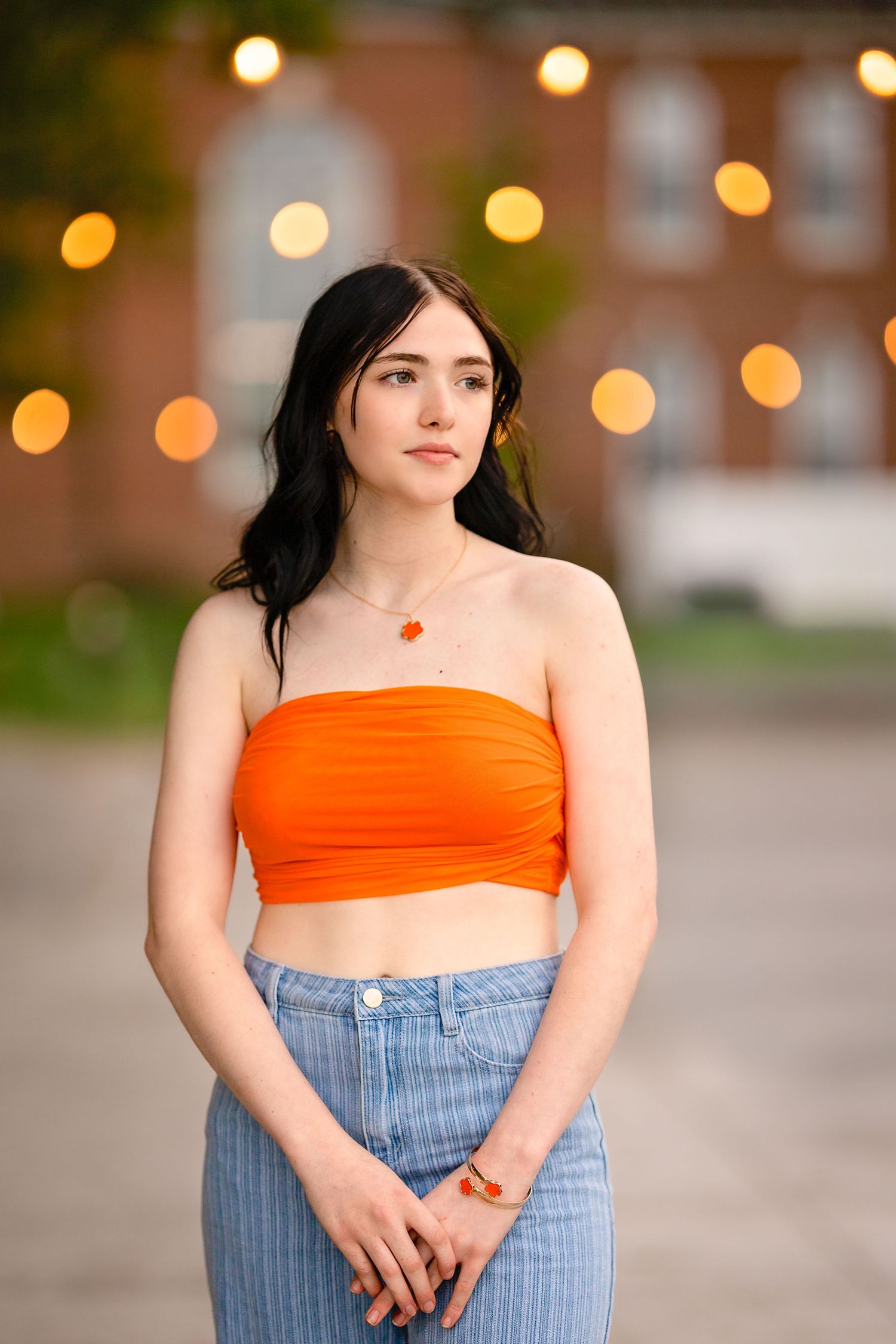 Portrait against evening bokeh lights featuring an orange crop top and high-waisted jeans.