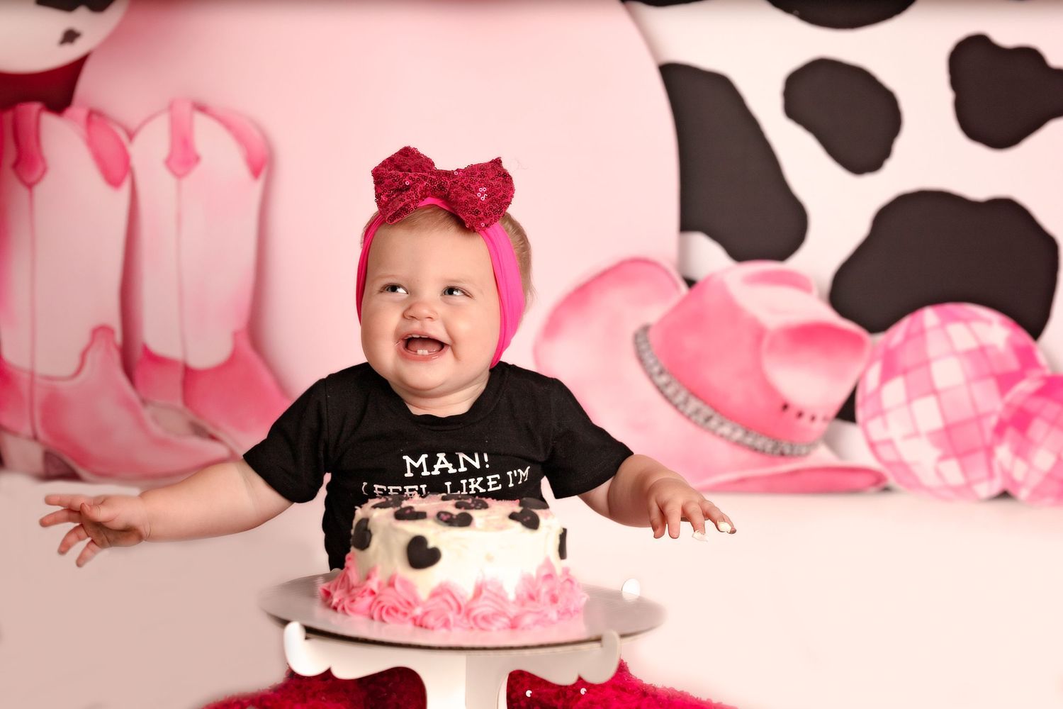 Baby wearing black shirt and pink bow celebrating first birthday with smashed cake against pink and cow print backdrop.