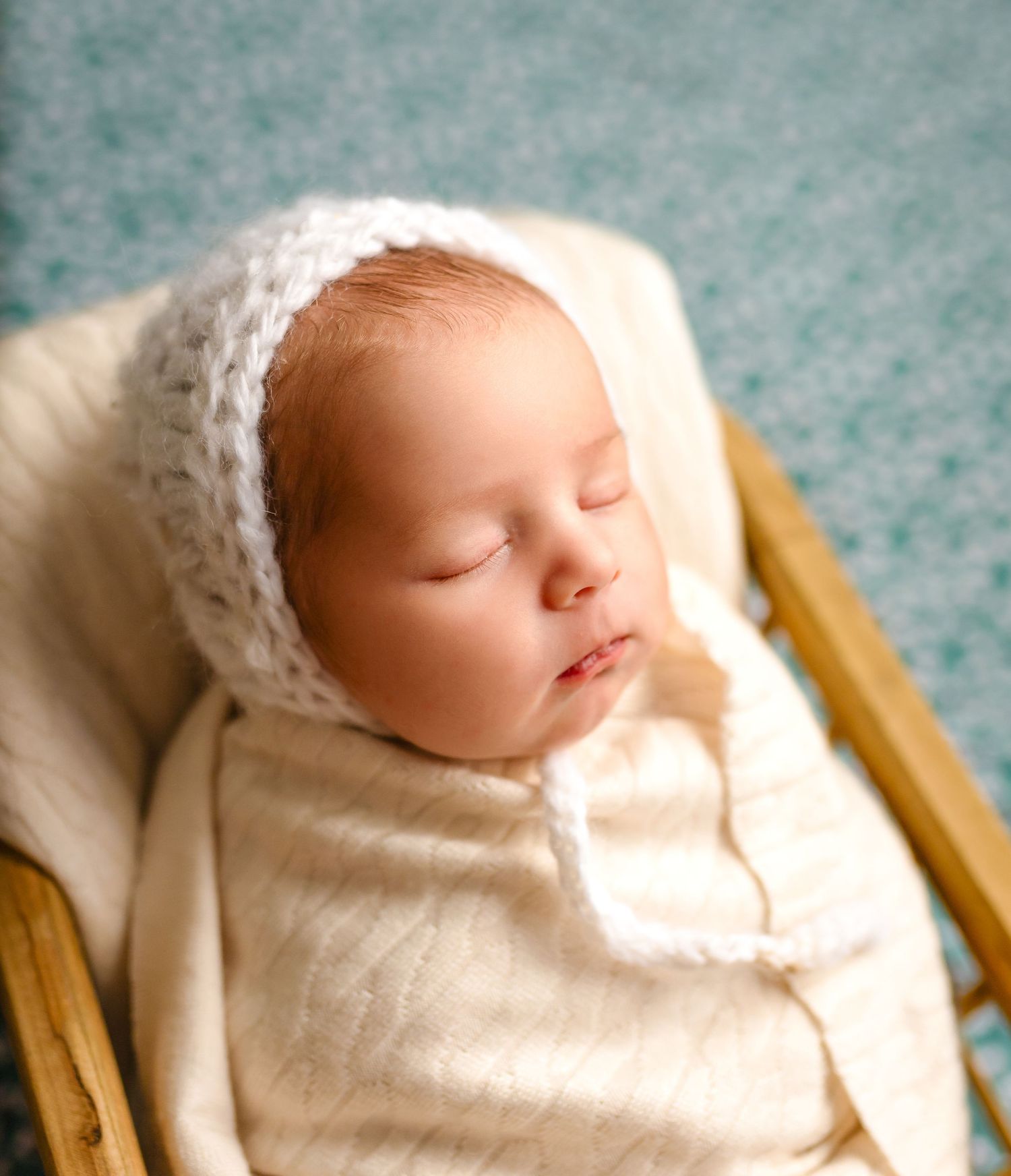 Sleeping newborn wearing soft white knitted bonnet and wrap in wooden bassinet.