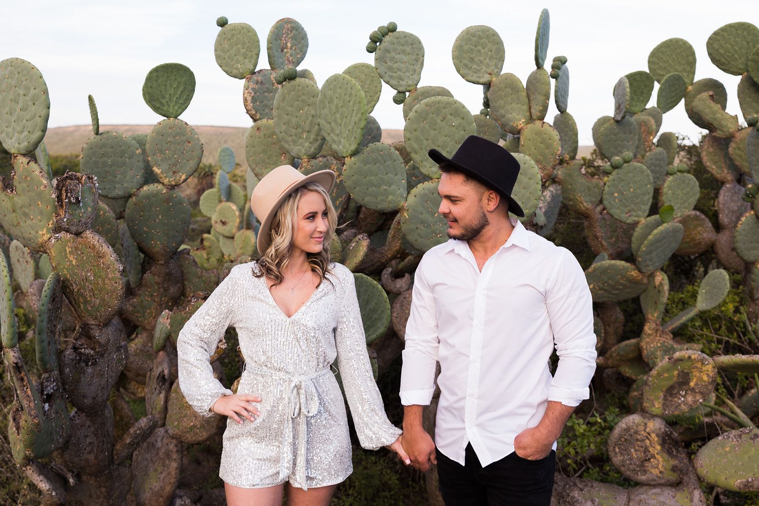 A couple poses together in white clothing for engagement photos among desert cactus and succulents at sunset.