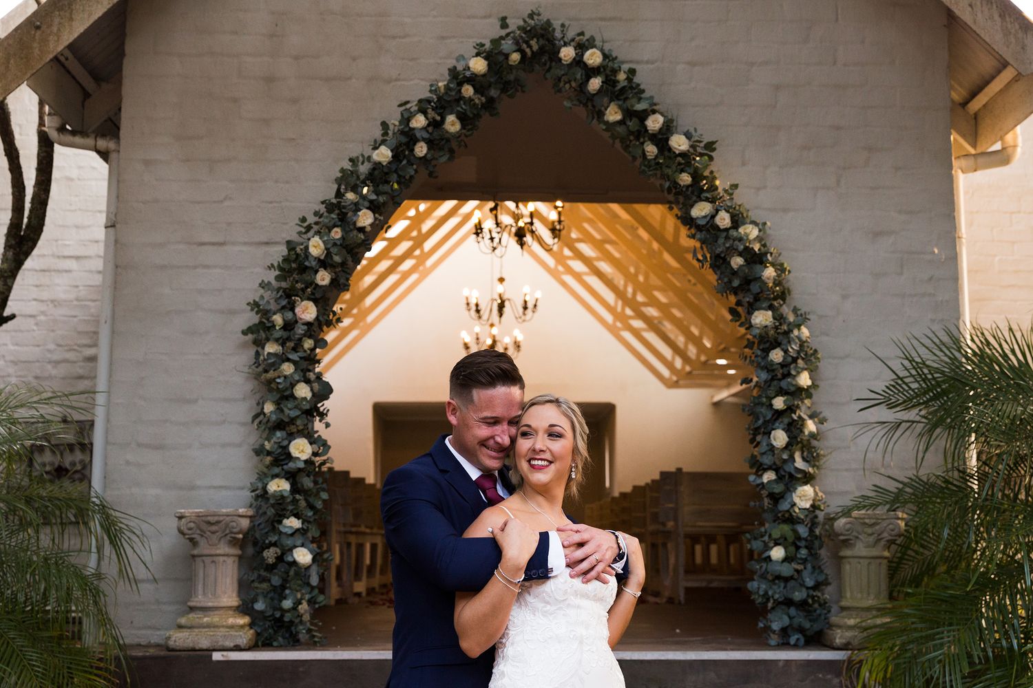 A couple embraces under evergreen holiday archways decorated with twinkling lights in an elegant indoor setting.