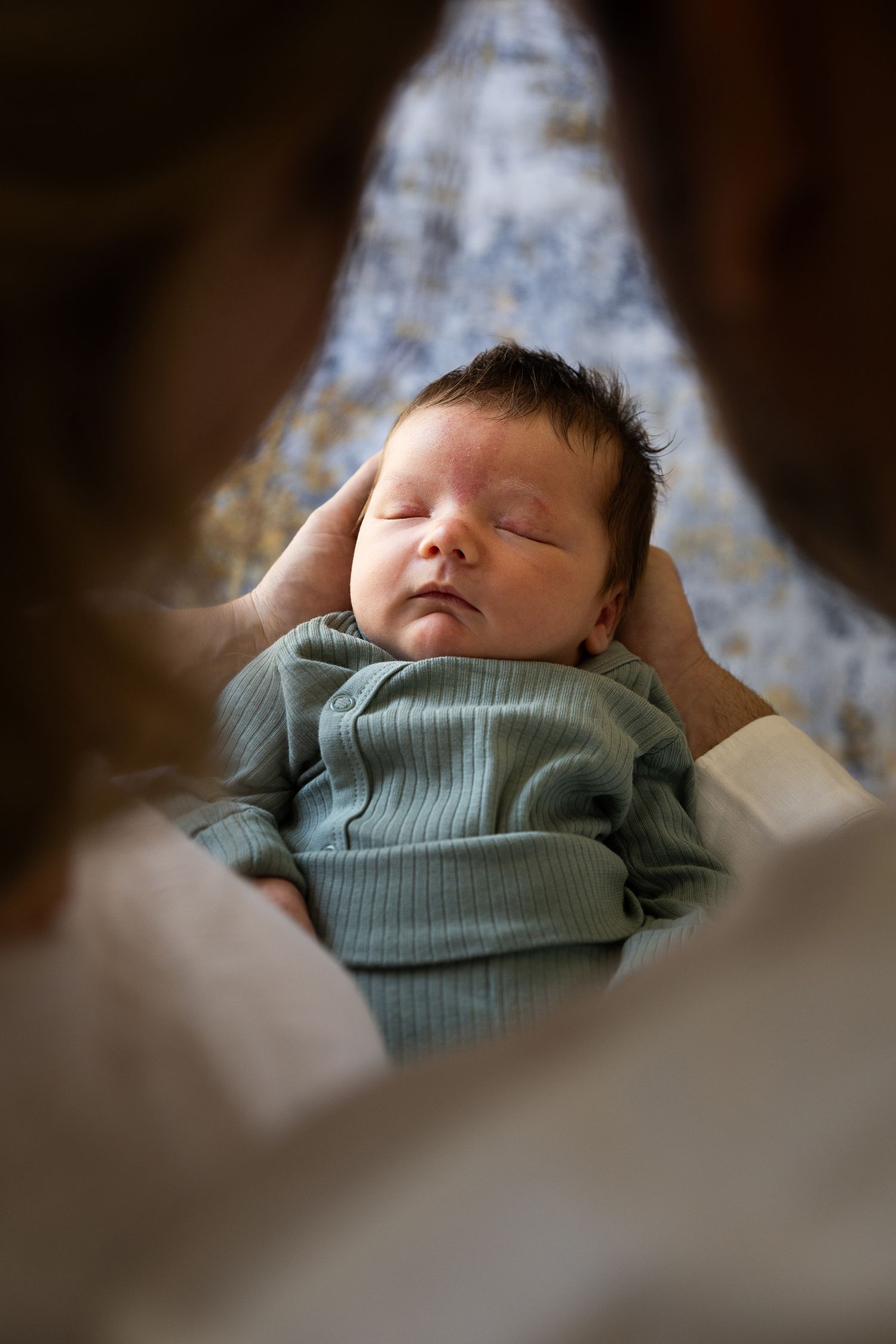 A sleeping newborn wearing a soft mint green sweater is photographed in a cozy indoor setting.