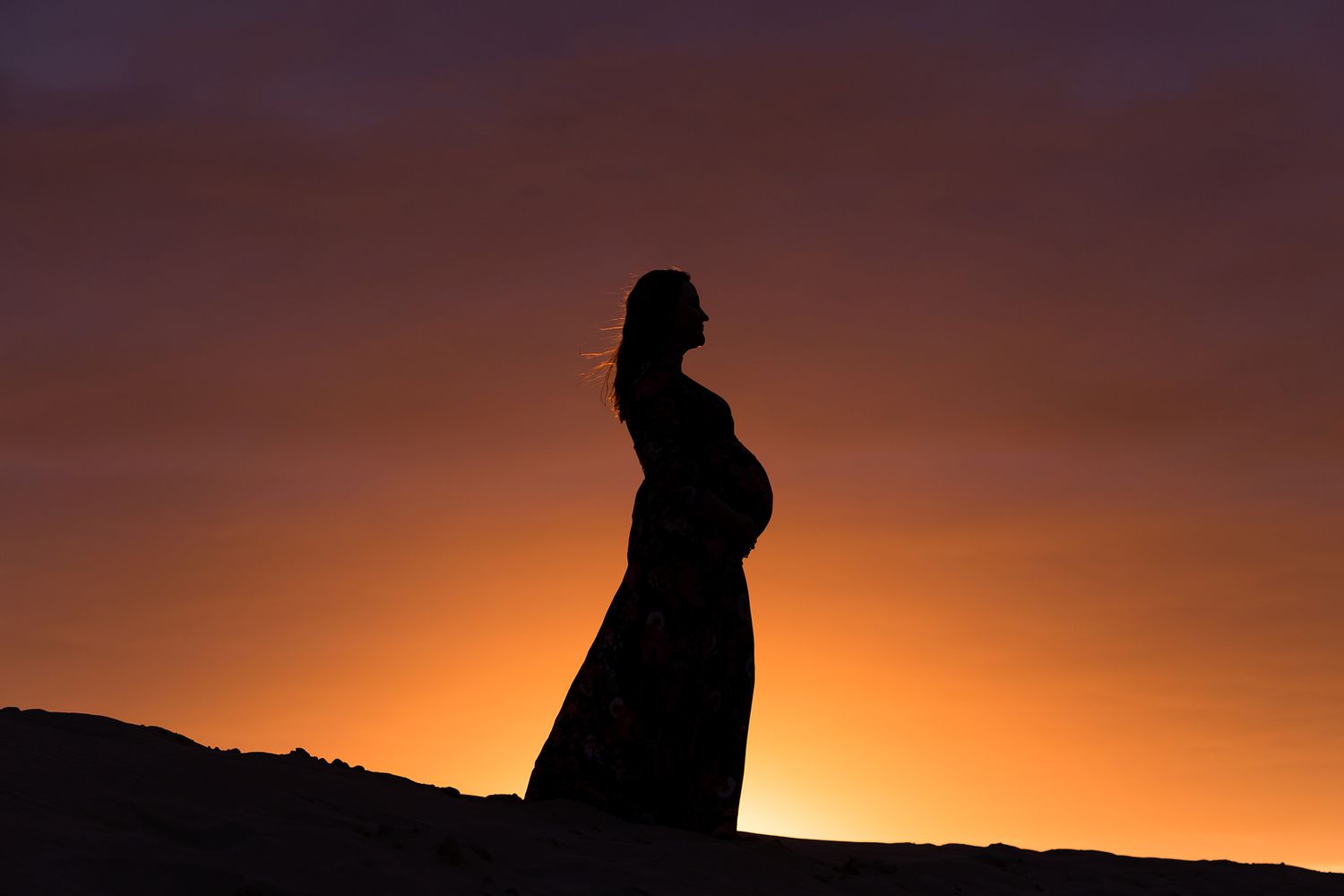 Silhouette of a pregnant woman in a flowing dress against a vibrant orange sunset sky.
