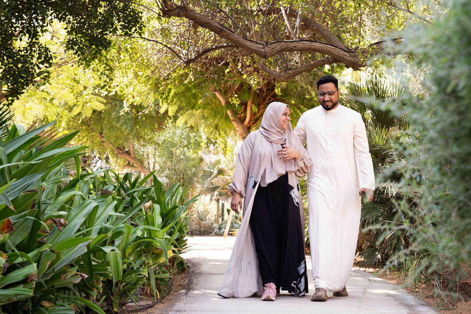 Couple takes romantic stroll along tree-lined pathway during sunny outdoor photoshoot.