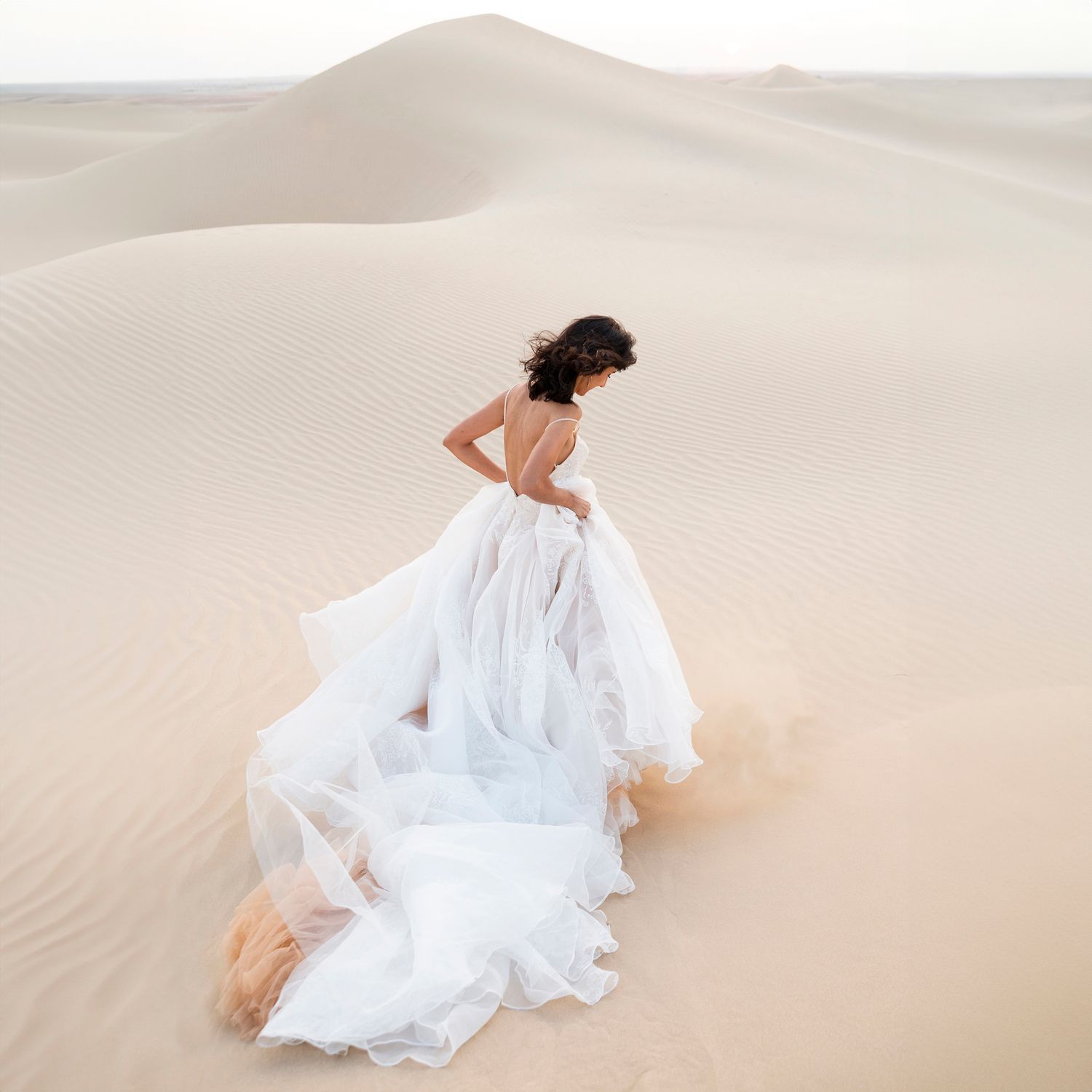 Woman in flowing white dress poses gracefully across desert sand dunes in ethereal bridal photoshoot.