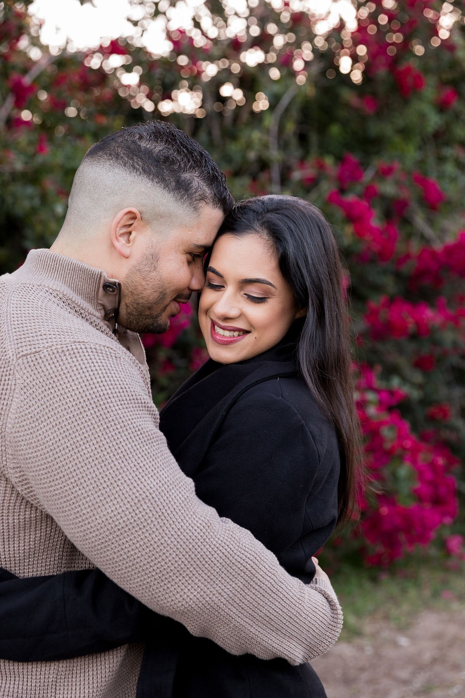 A romantic couple embraces in front of vibrant red bougainvillea flowers on a sunny afternoon.