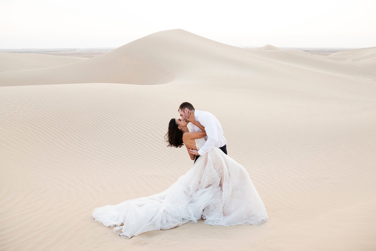 A couple in wedding attire embraces romantically while sitting on pristine white sand dunes in a desert landscape.
