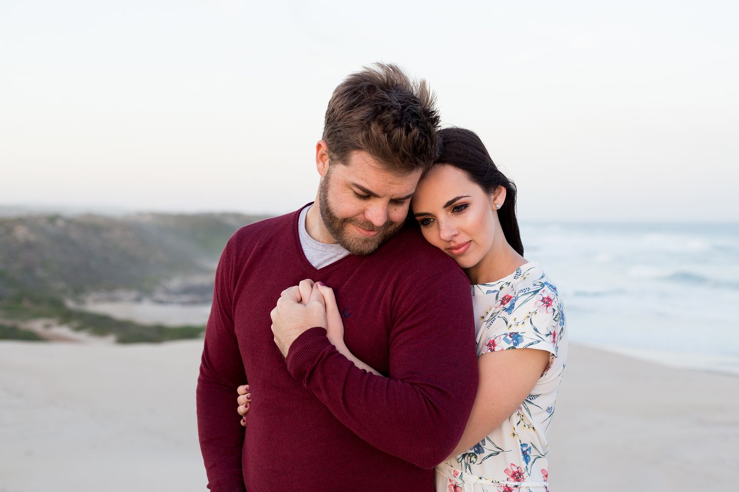 Romantic beach photoshoot shows couple in burgundy and floral outfits embracing by the ocean.