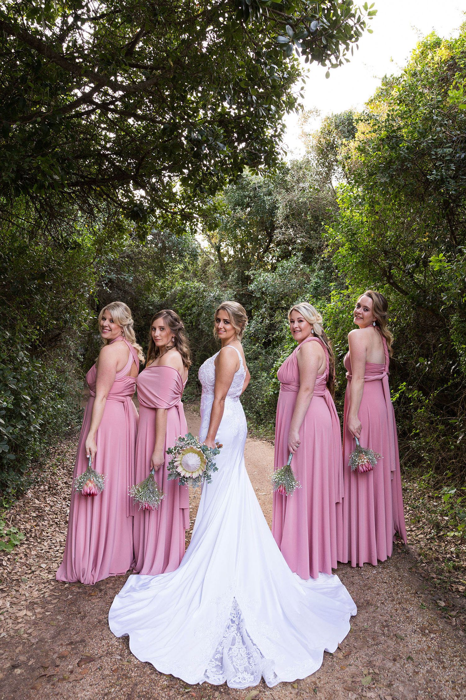 A bridal party in dusty rose pink dresses and white wedding gown poses together on a wooded path.