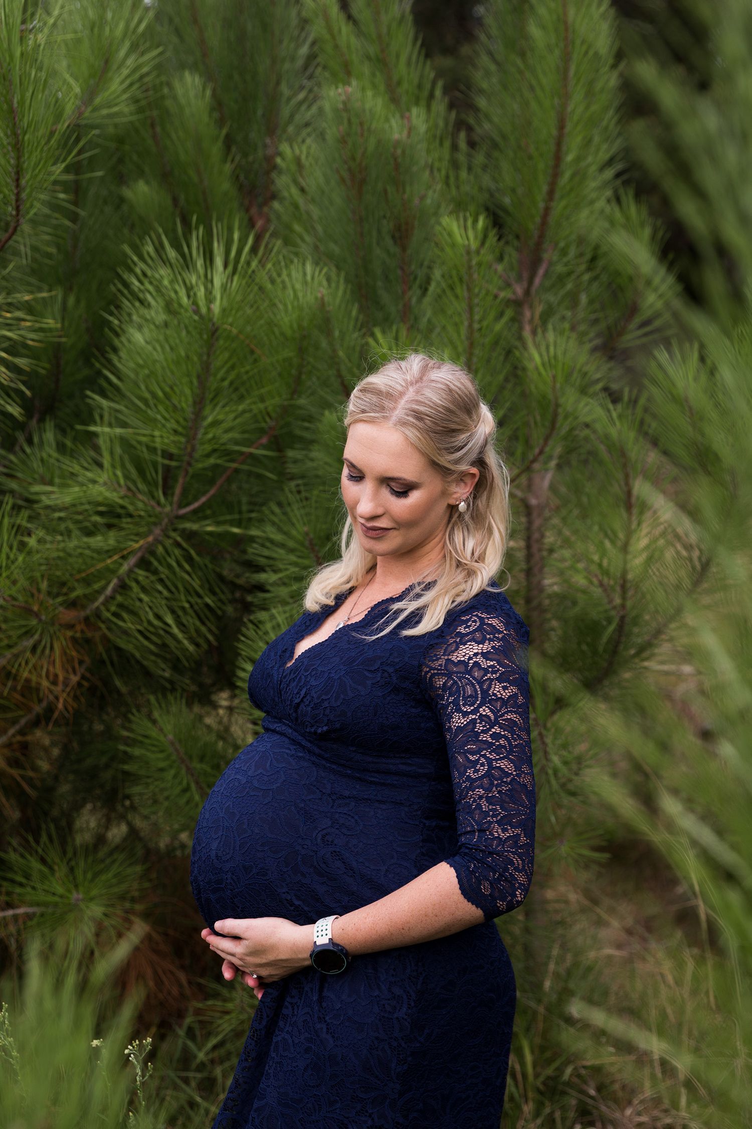 A pregnant woman in a navy lace dress poses against a backdrop of green pine trees.