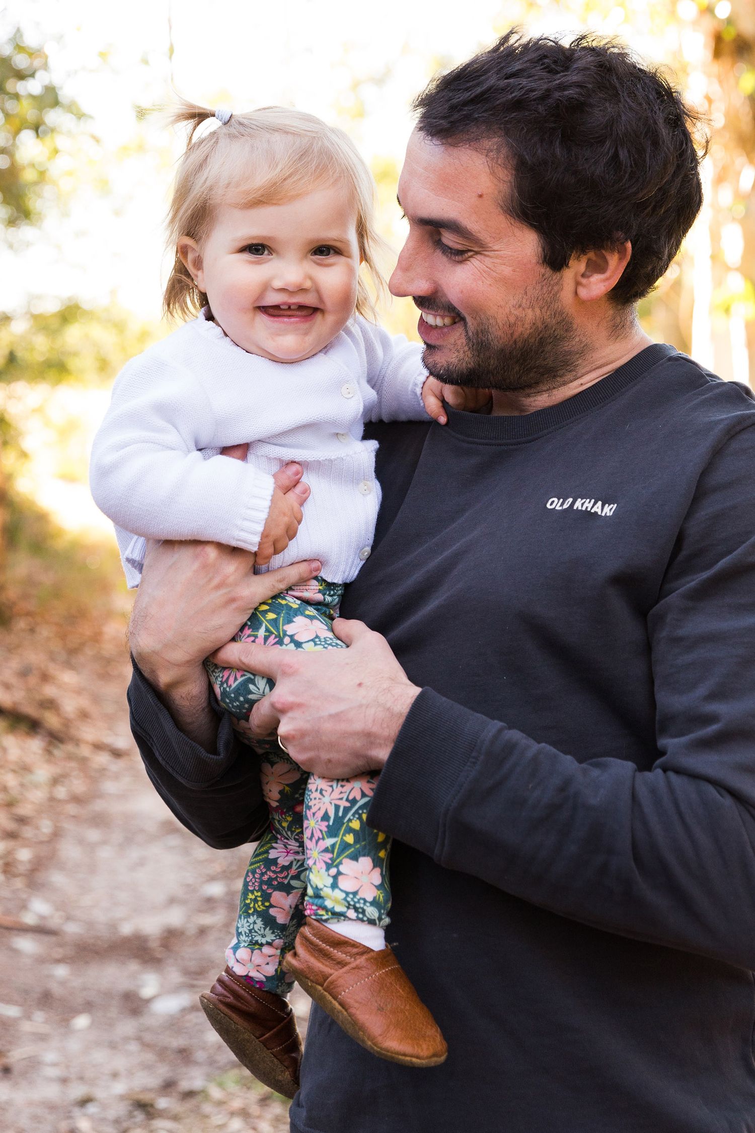 Parent in black sweater holding a smiling toddler wearing floral pants against a sunlit outdoor background.