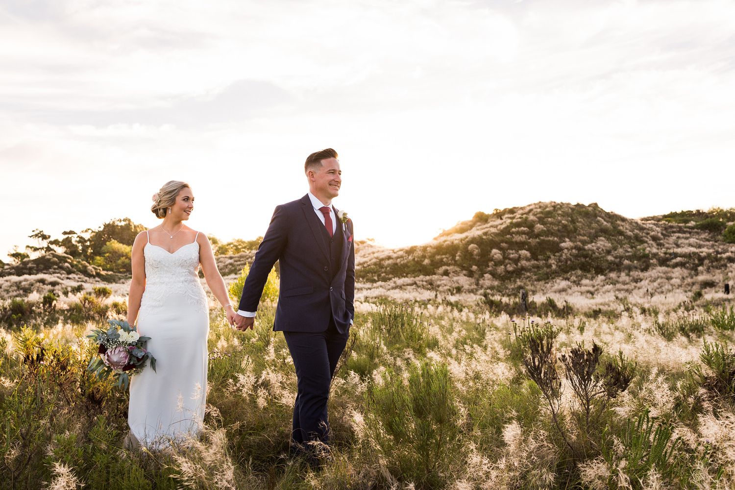 A couple in wedding attire walks hand in hand through a scenic coastal dune landscape at golden hour.