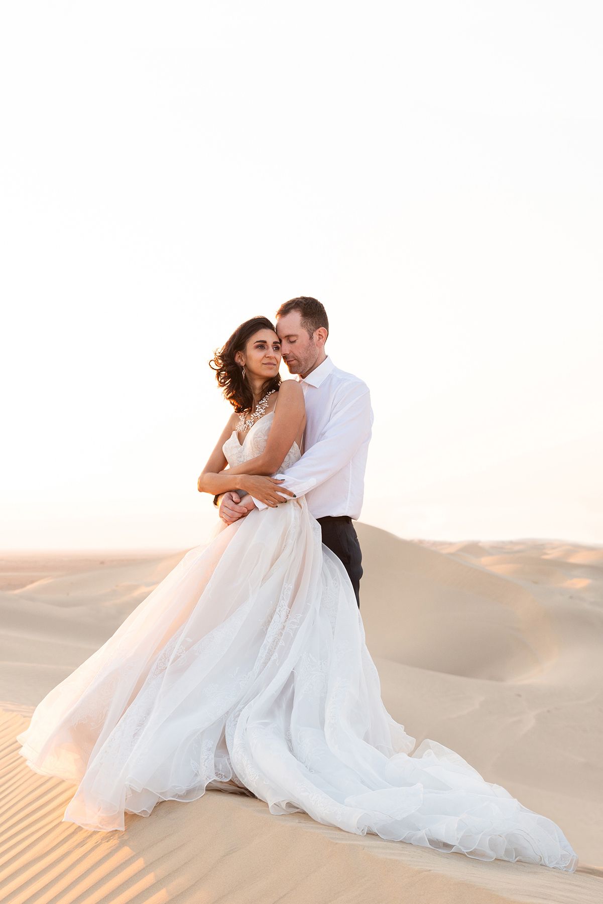 A romantic wedding photoshoot in white sand dunes at sunset shows tender moments between newlyweds.