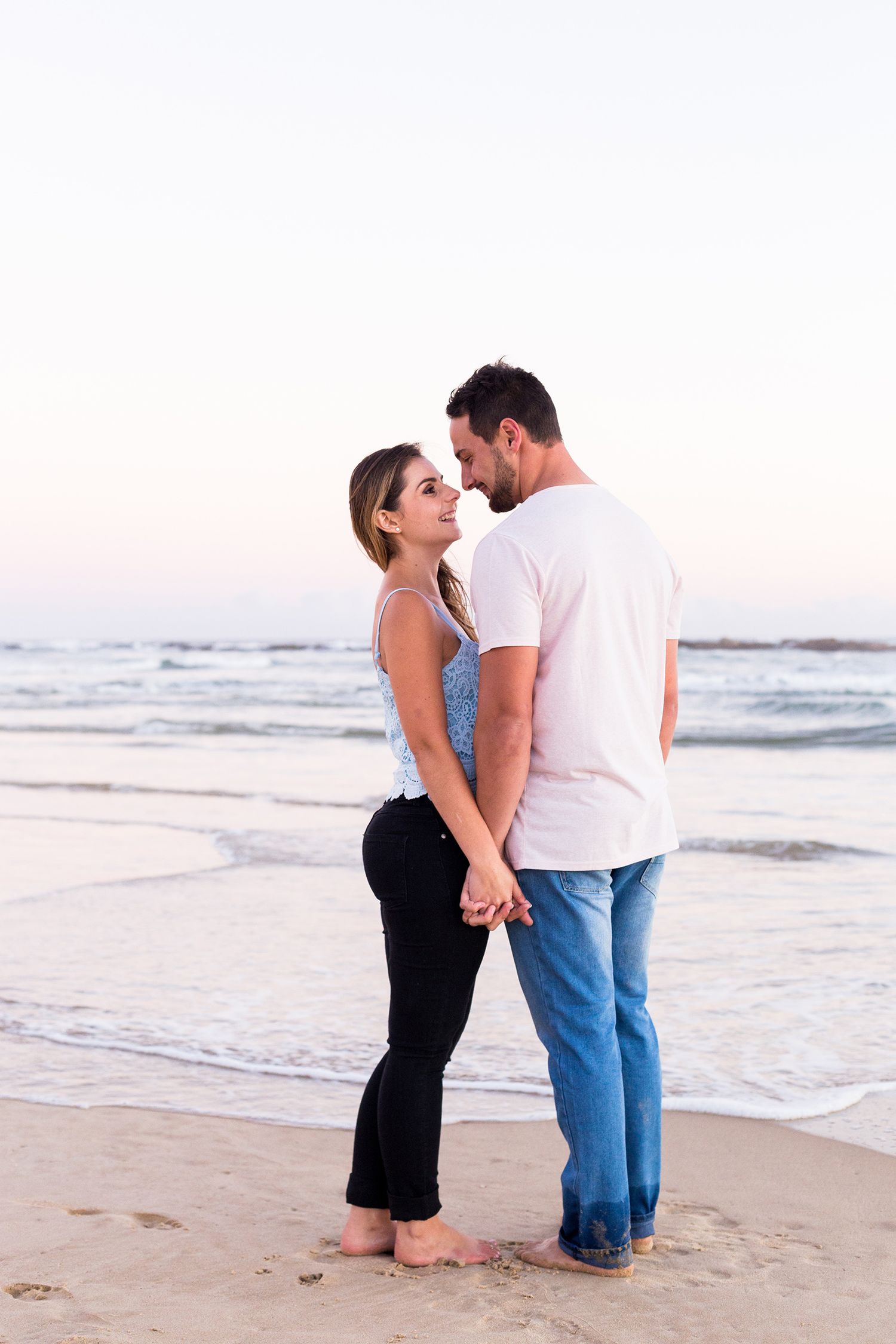 A romantic couple shares an intimate moment while standing barefoot on a beach at sunset.