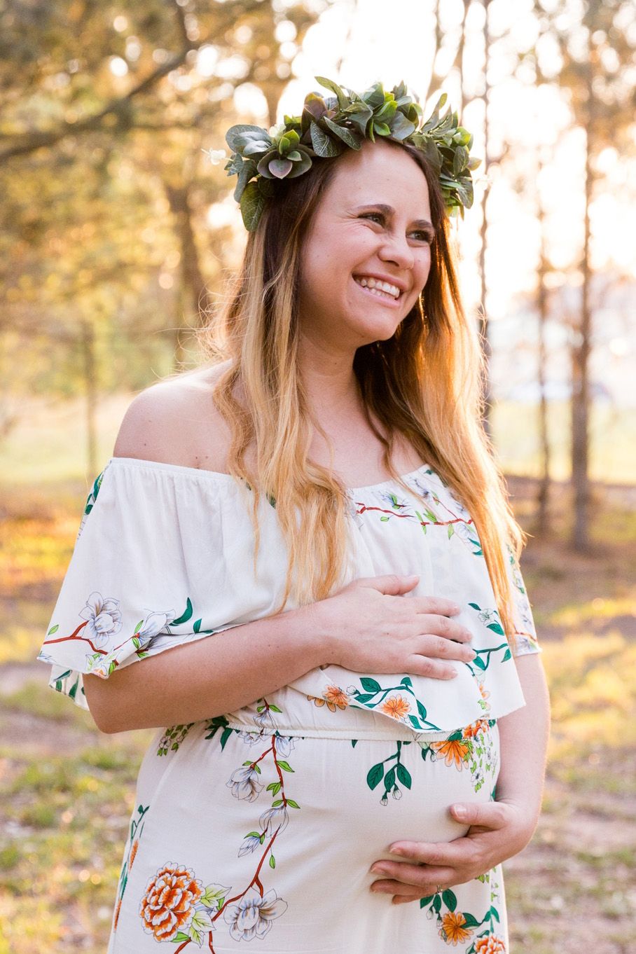 A pregnant woman in a floral dress and flower crown poses outdoors during golden hour in a wooded setting.