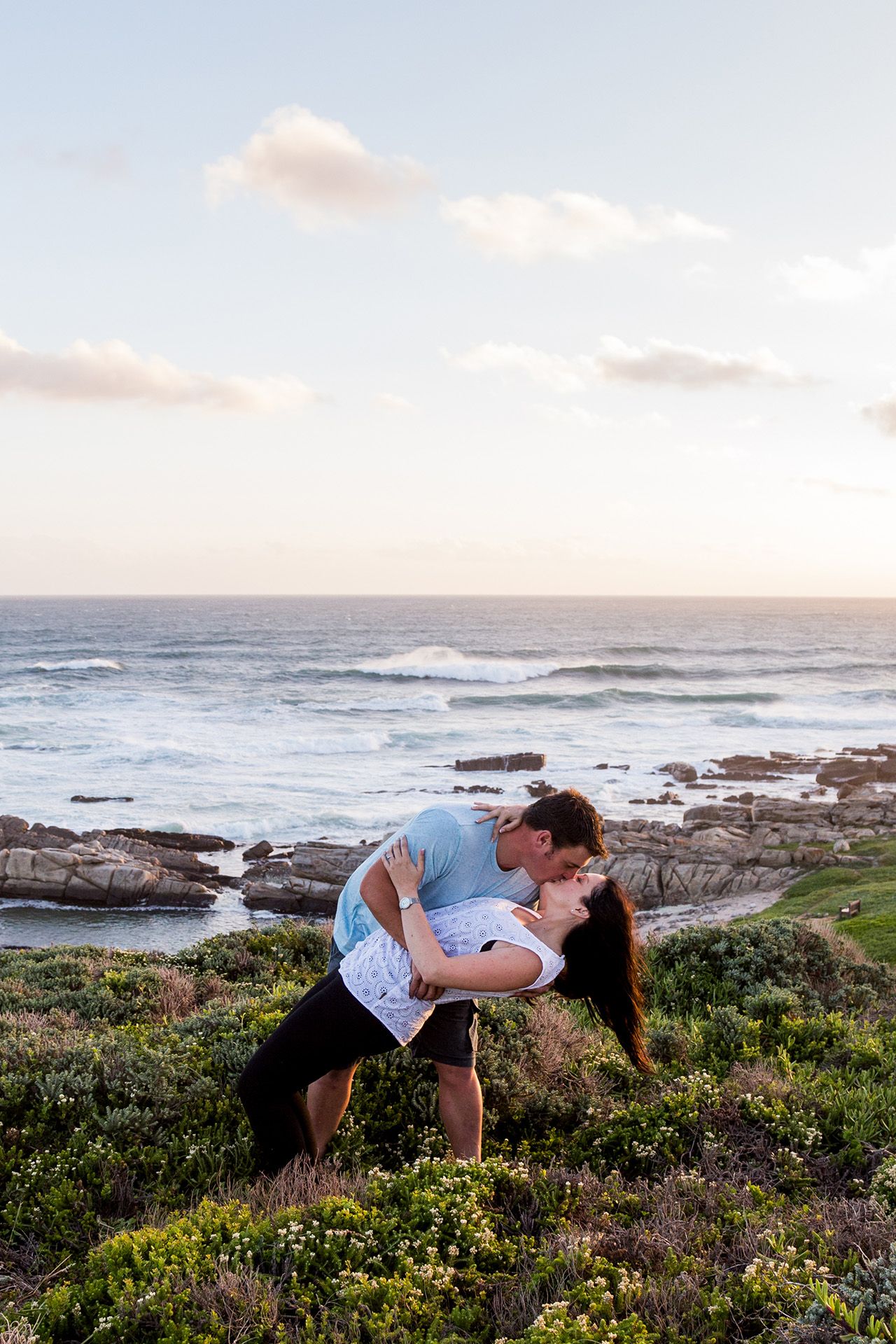 Two people share a romantic moment on a rocky coastal cliff overlooking the ocean at sunset.