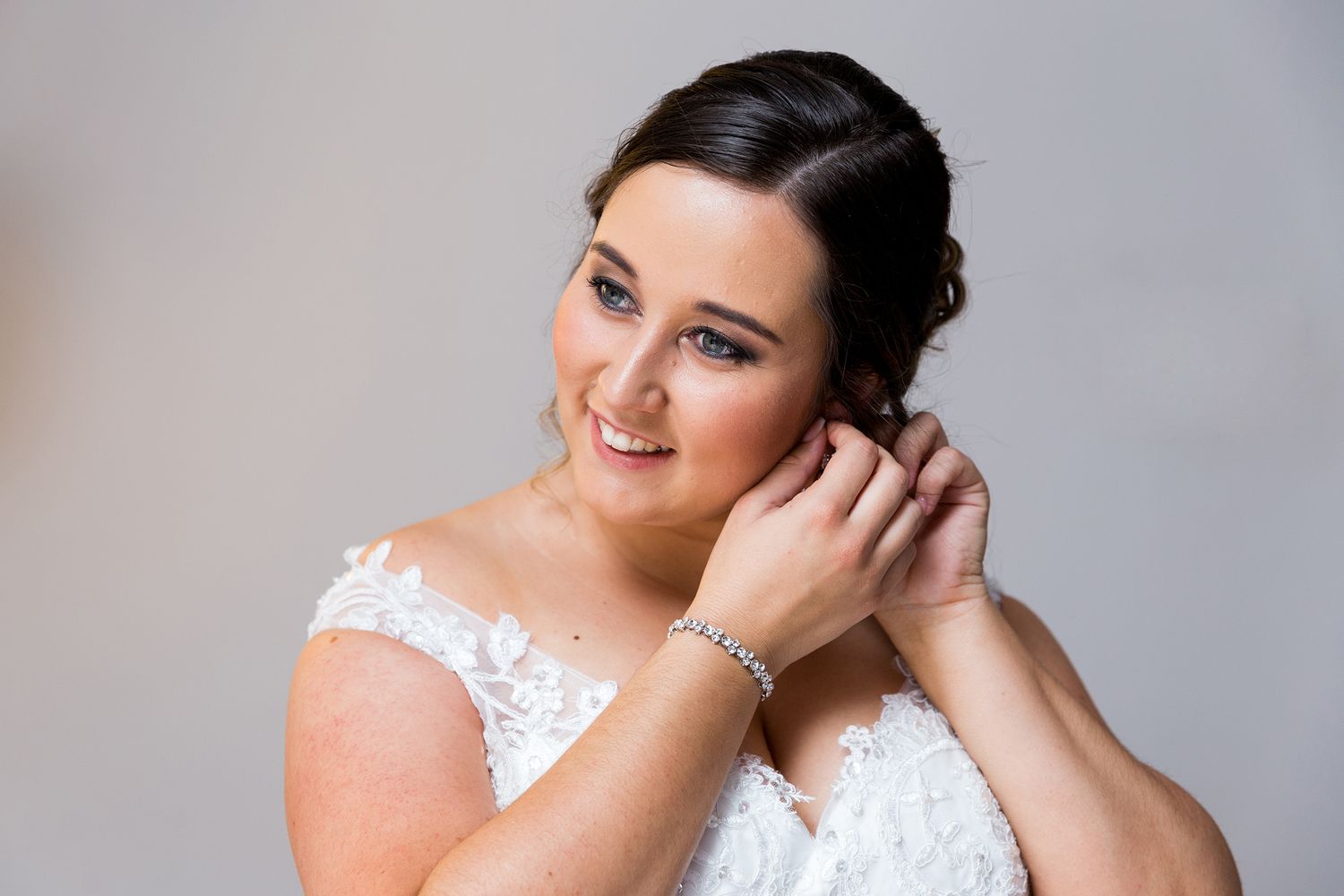A bride wearing a white lace wedding dress and silver jewelry touches her hair while posing on a light background.