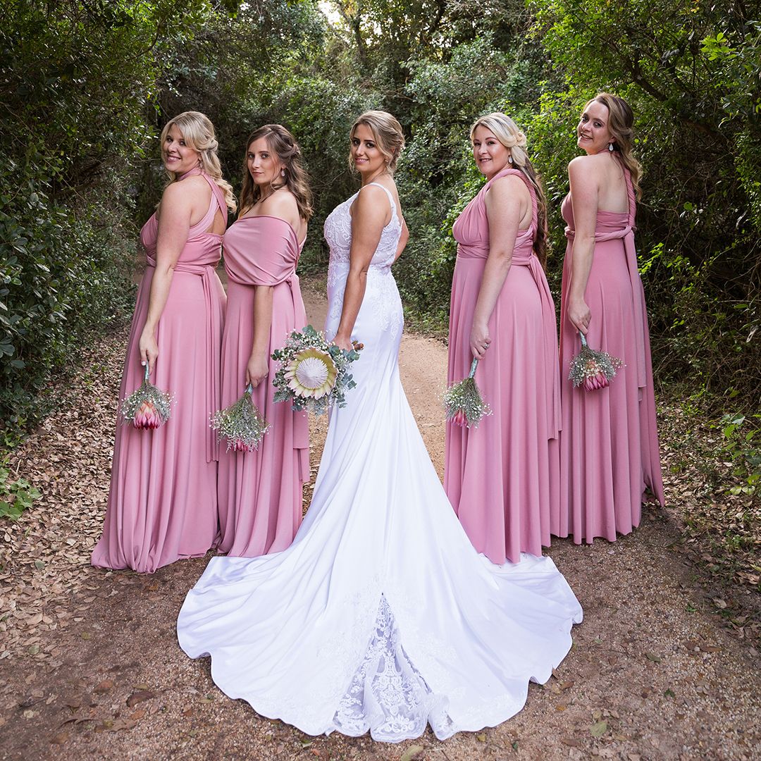 A bride in white and bridesmaids in dusty rose dresses pose together on a tree-lined outdoor path during a wedding.