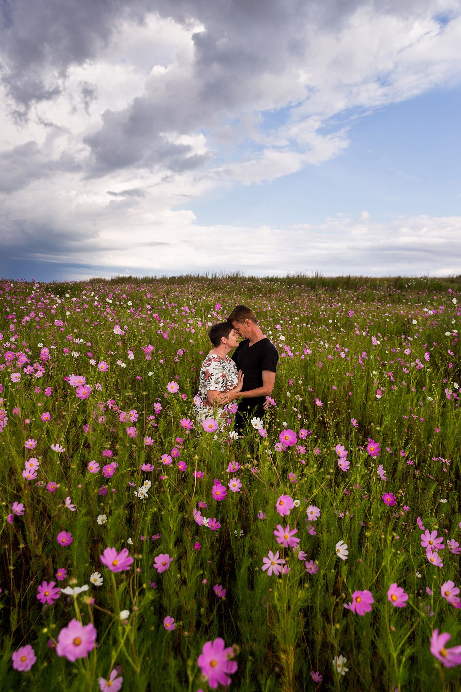Couple embraces in a beautiful meadow filled with blooming pink wildflowers under dramatic cloudy skies.
