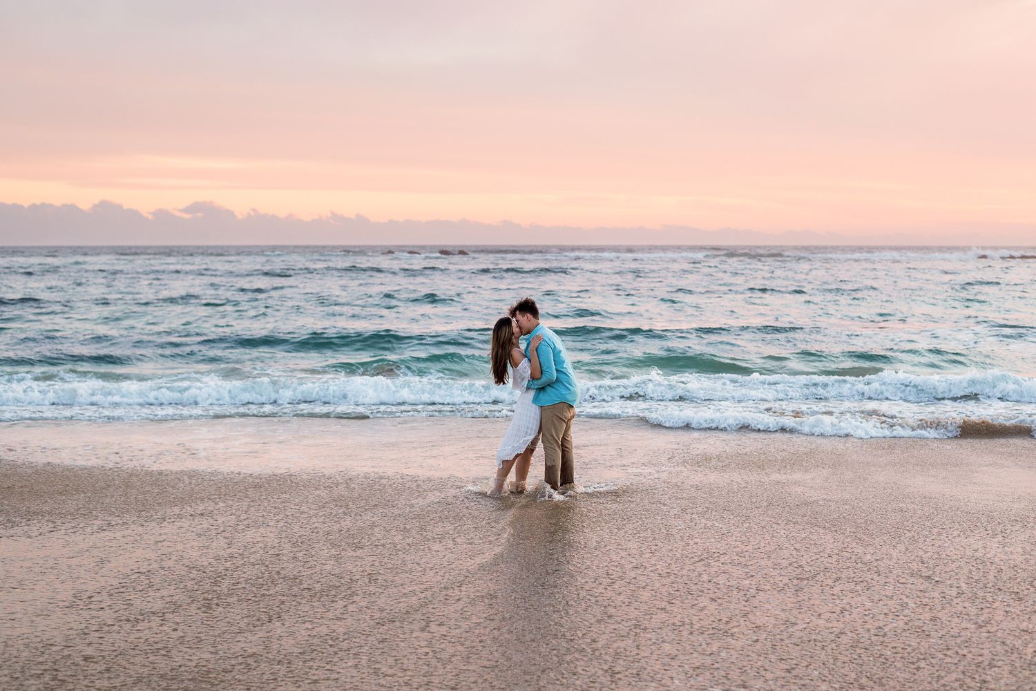 Couple embracing on sandy beach at sunset with pink-orange sky and rolling ocean waves in the background.