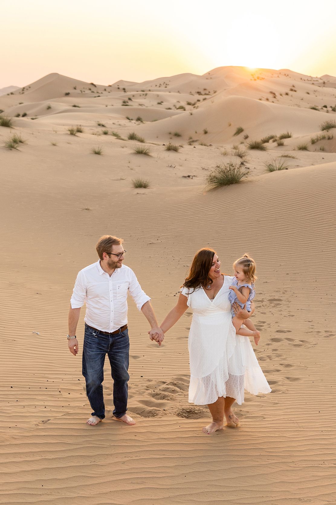 Family walking together through sandy desert dunes at sunset with soft golden lighting creating long shadows.