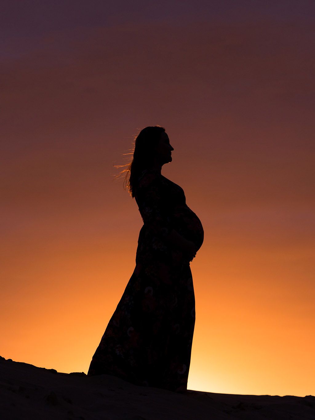 Silhouette of a pregnant woman cradling her belly against a vibrant orange and purple sunset sky.