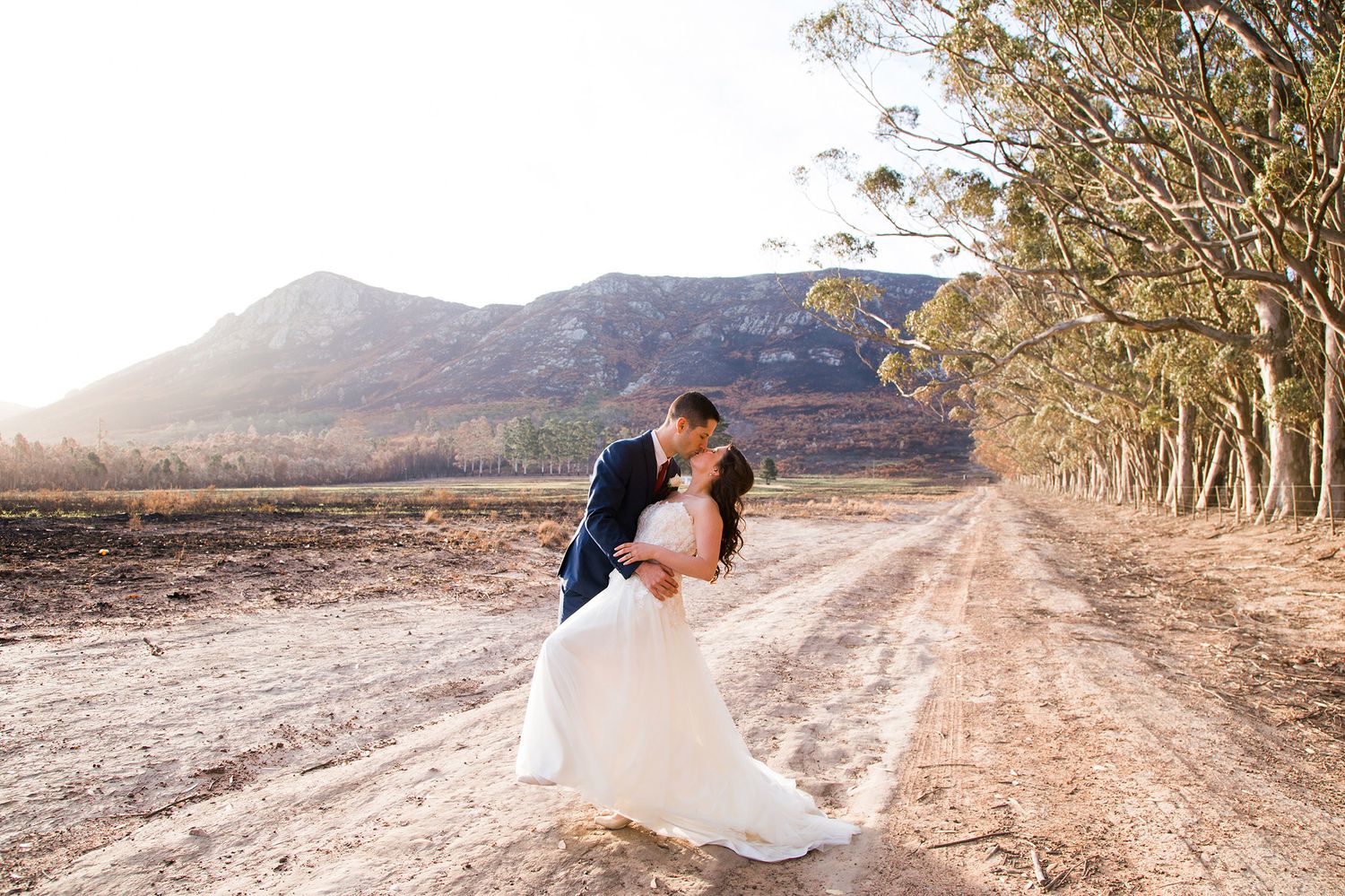A romantic couple embracing and kissing on a dusty mountain road during golden hour in nature.