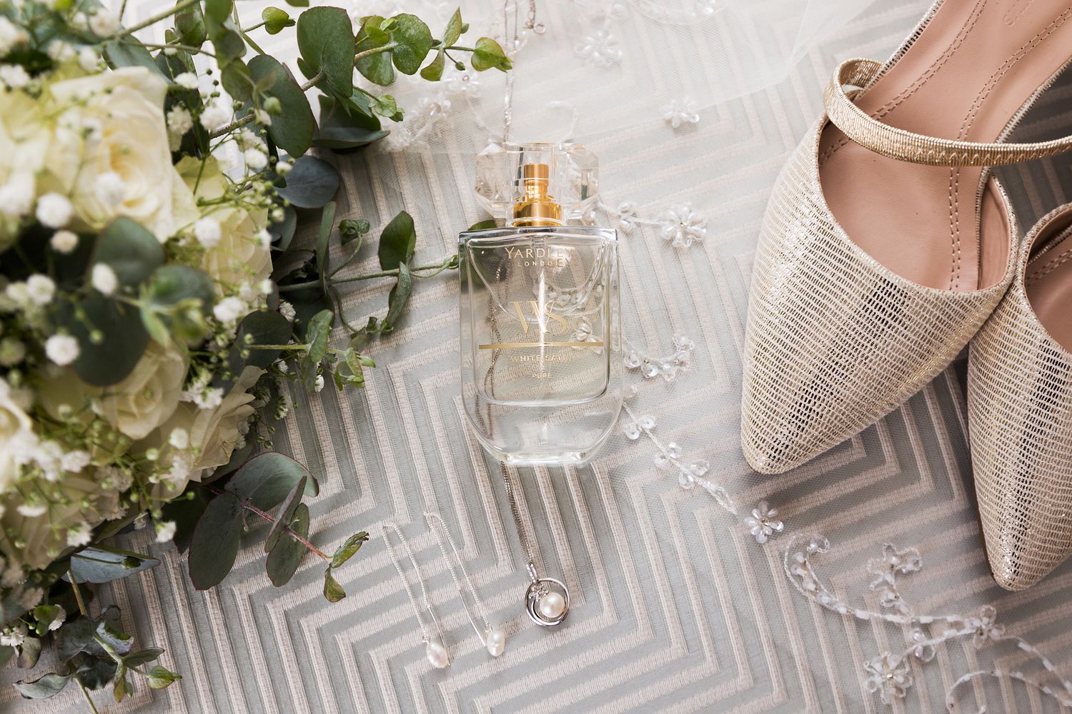 Wedding accessories including sparkly shoes, white florals, perfume bottles and chevron fabric arranged in an elegant layout.