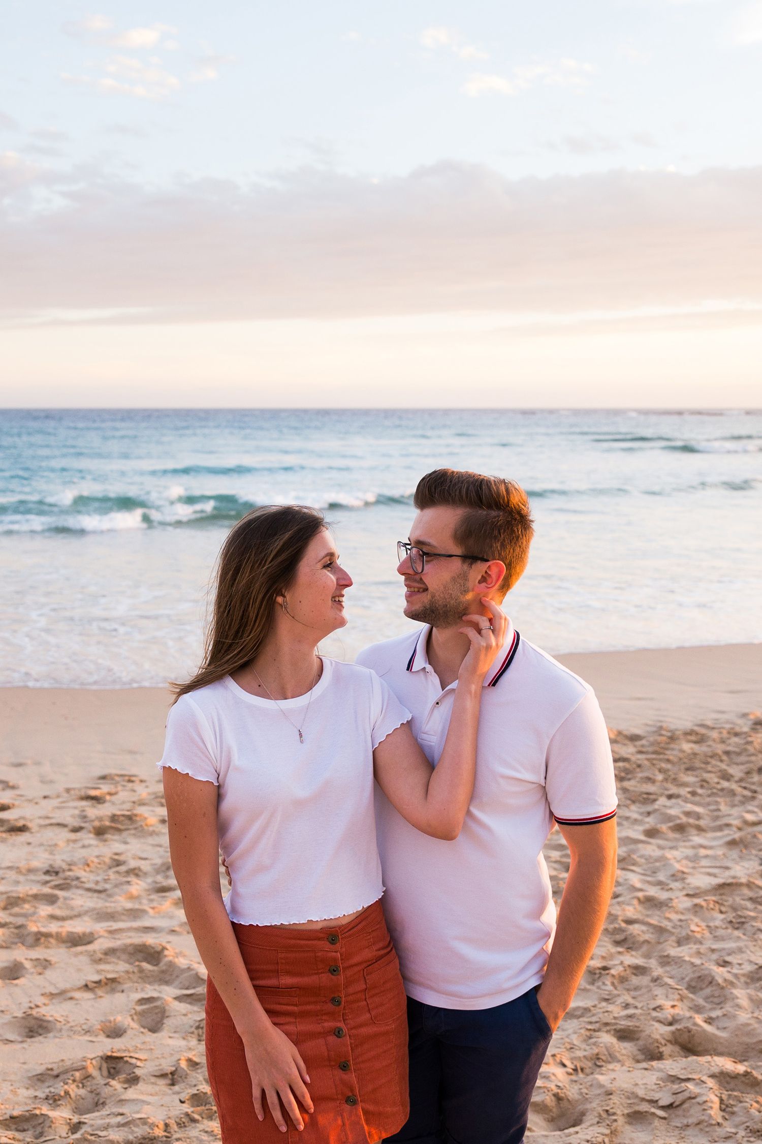 A couple enjoys a romantic sunset moment together while sitting on a sandy beach with ocean waves in the background.