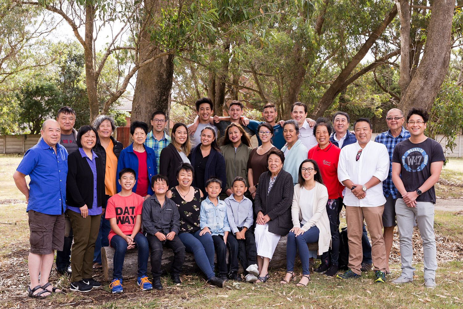 Large outdoor group portrait of people gathered under a tree in casual clothing during a sunny day event.