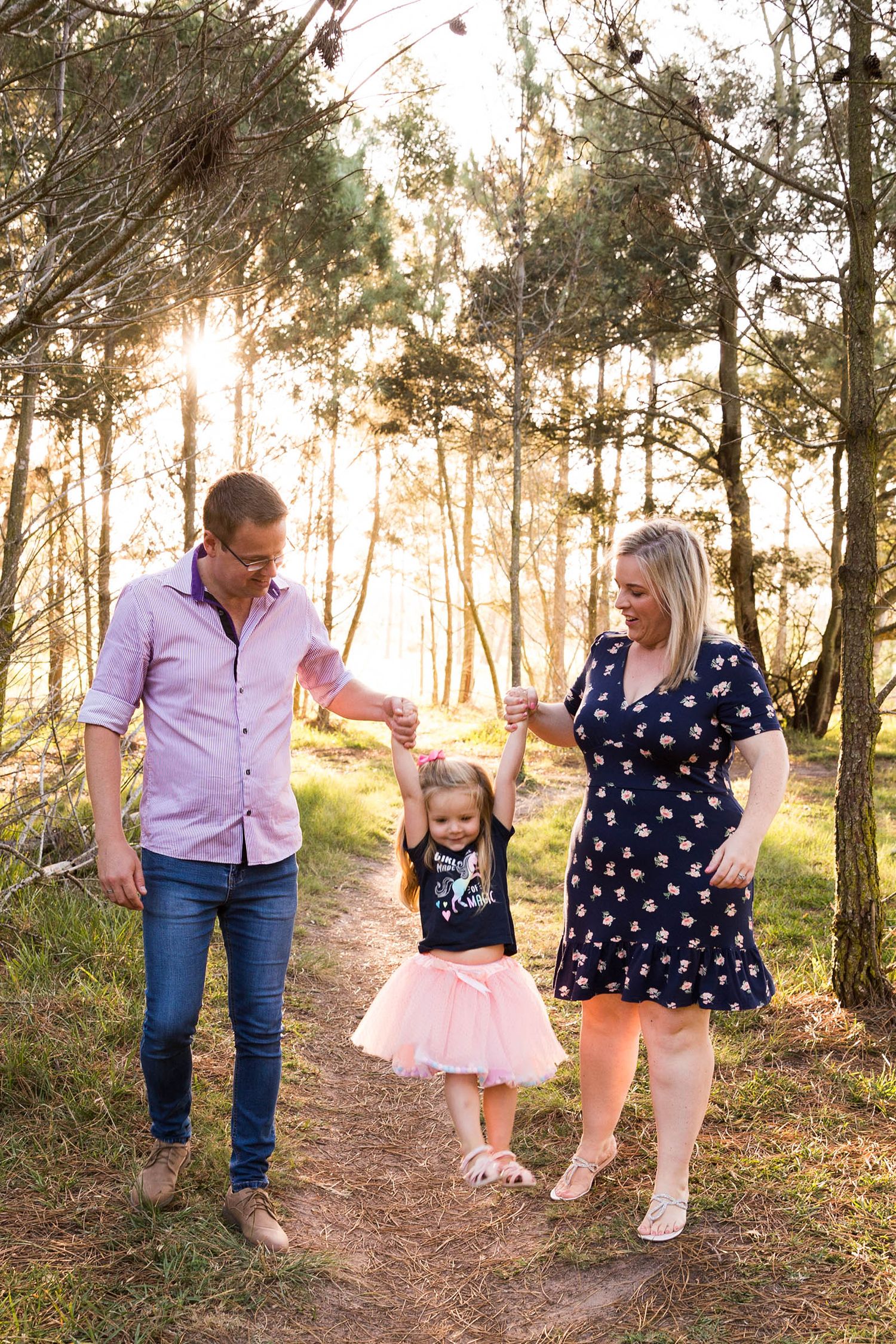 Family walking together on a wooded trail during golden hour with the sun filtering through pine trees.