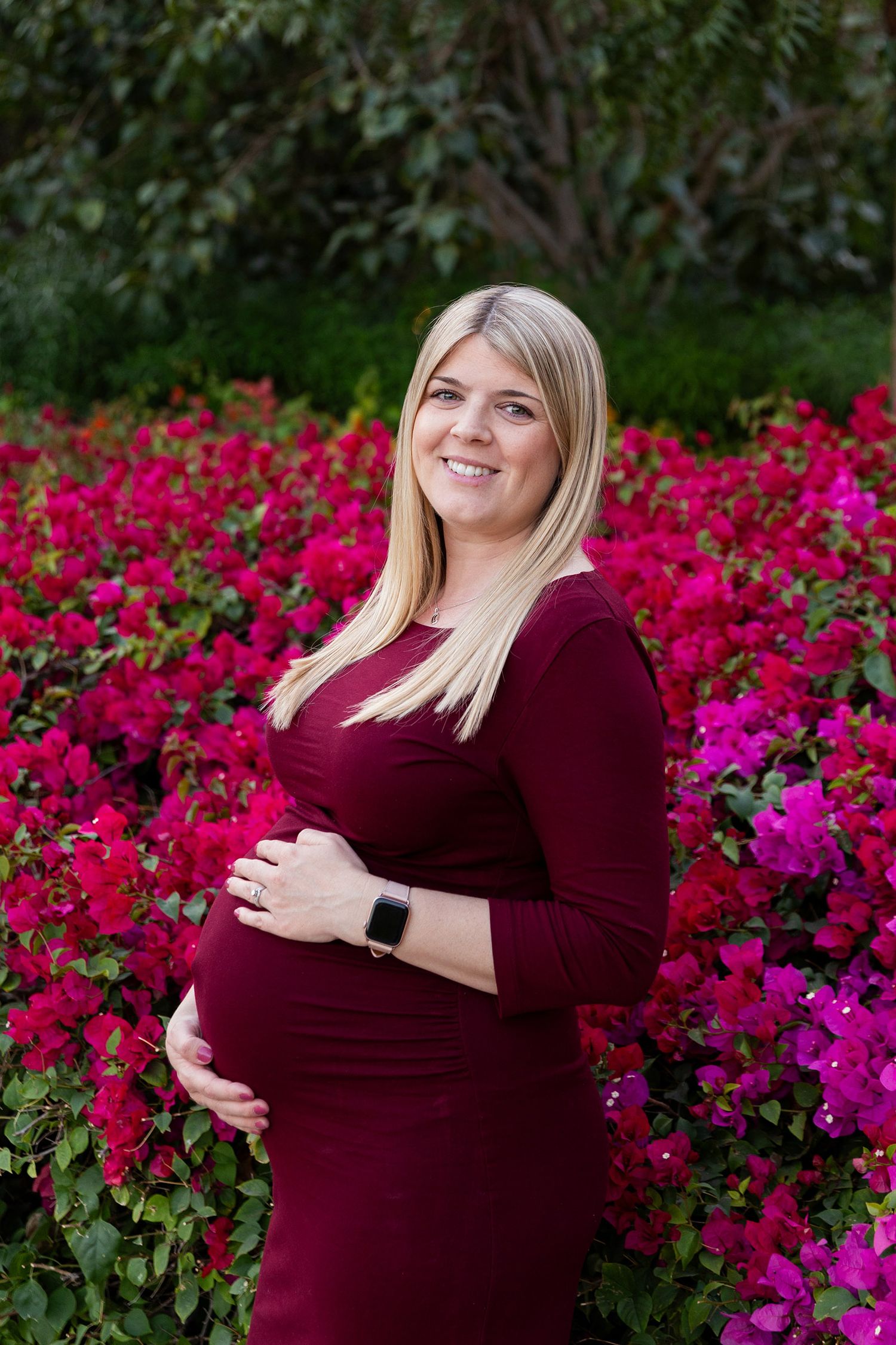 Woman in burgundy dress stands in front of vibrant pink flowers during maternity photoshoot outdoors.