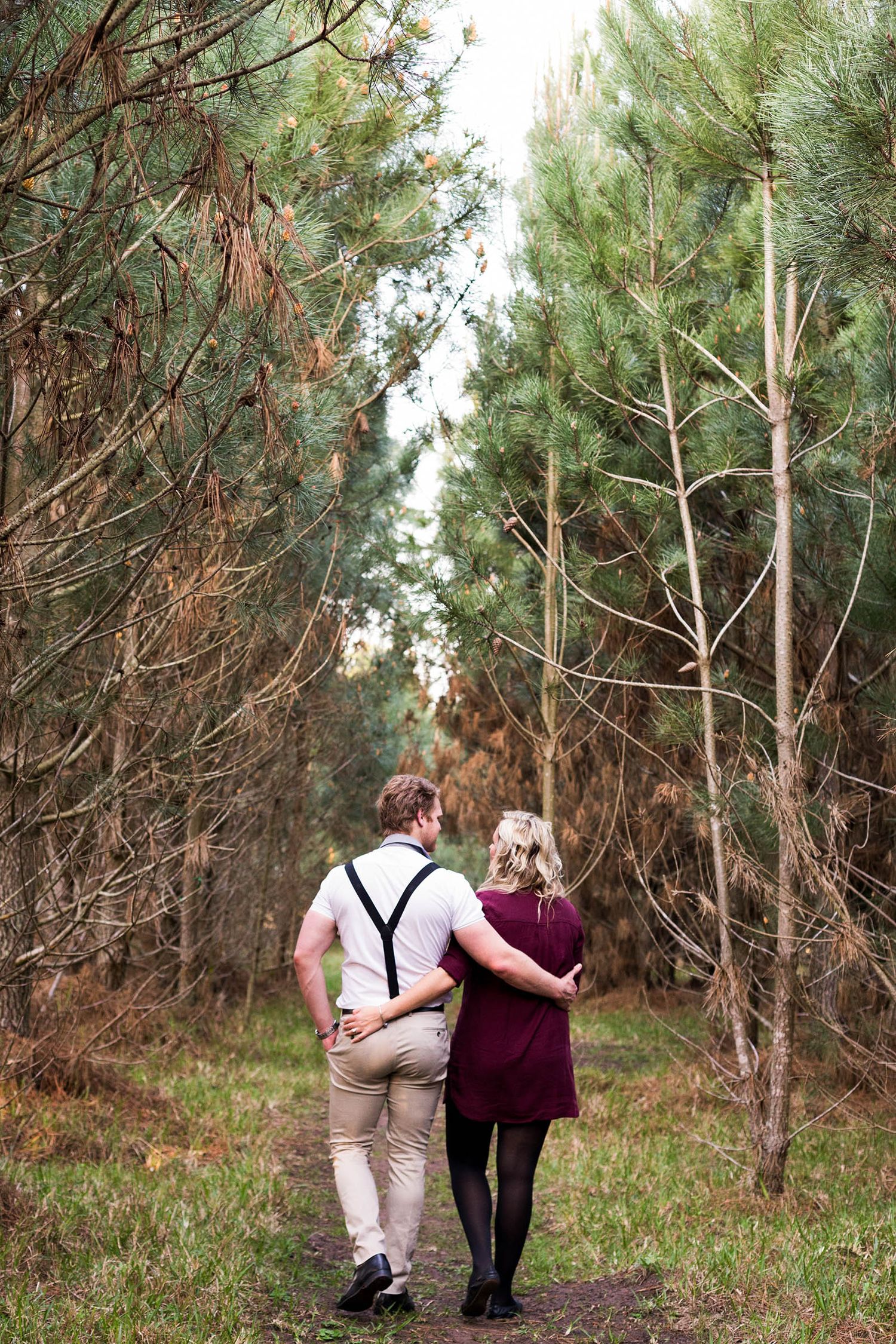Two figures walking hand in hand along a peaceful pine forest trail during autumn.