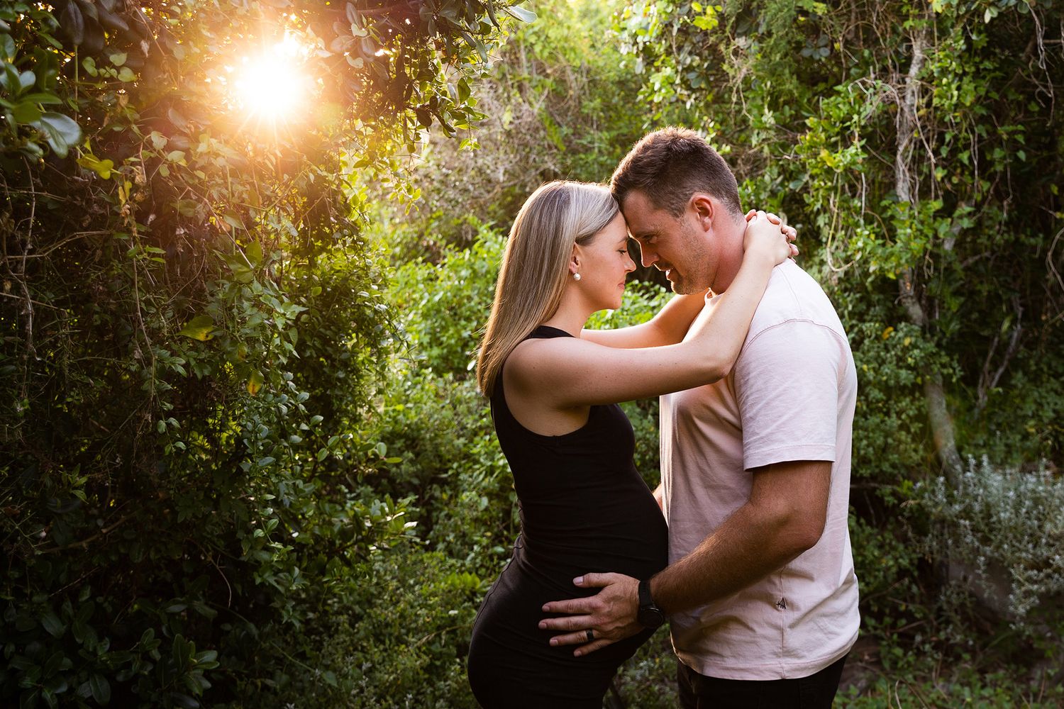 A couple shares tender moments in a garden setting during golden hour with sunlight filtering through lush greenery.